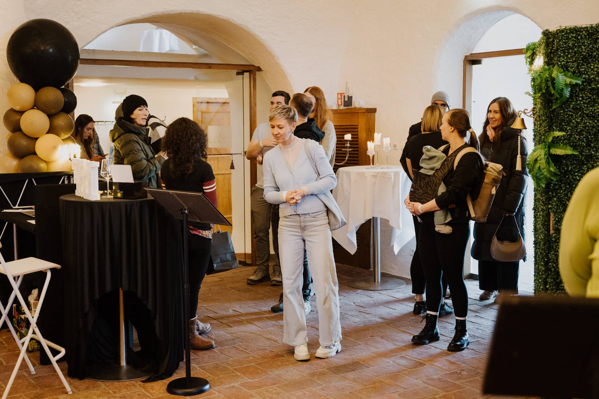 Eine Gruppe von Menschen trifft sich drinnen bei einer Veranstaltung mit Backsteinböden, großen Fenstern und grüner Dekoration. Einige Gäste stehen in der Nähe eines Tisches mit schwarz-goldenen Luftballons, während andere sich in der Nähe des Eingangs unterhalten.