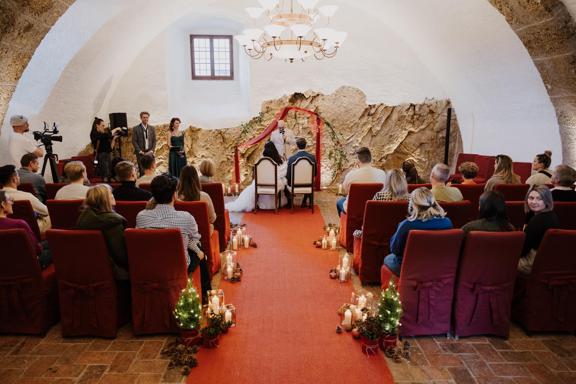 Eine Hochzeitszeremonie in einem rustikalen, gewölbten Raum mit roten Stühlen, einem mit Kerzen und kleinen geschmückten Bäumen gesäumten roten Teppich und Gästen, die sitzen, während das Paar unter einem Bogen aus grünem und rotem Stoff am Altar sitzt.