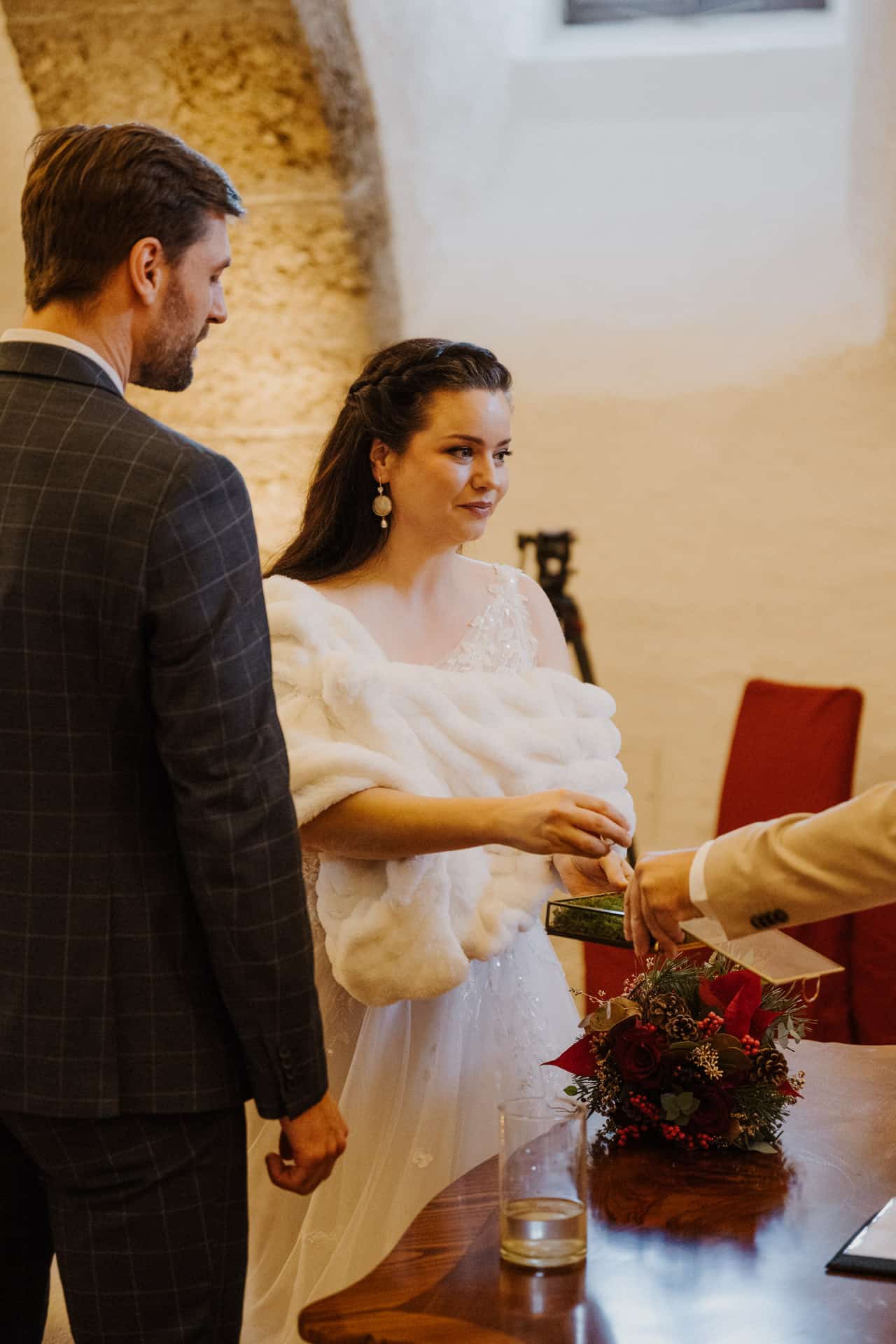 Eine Braut in einem weißen Kleid und einem Pelzschal steht neben einem Bräutigam in einem karierten Anzug. Sie reicht einer Person auf der anderen Seite eines Holztisches etwas, auf dem ein Blumenstrauß und ein Glas stehen. Die Umgebung scheint ein Innenraum mit Steinwänden zu sein.