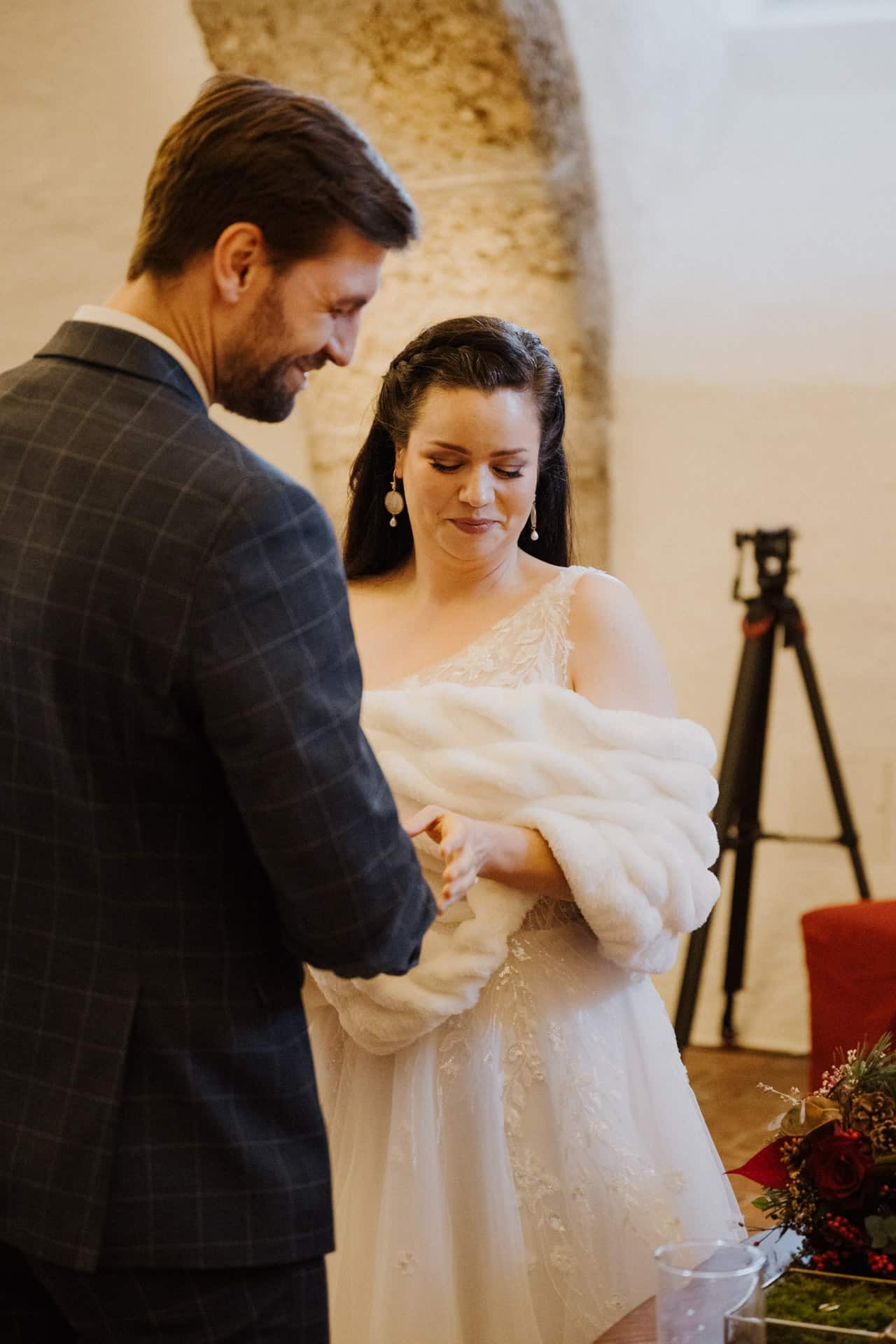 Eine Braut in einem weißen Kleid und einem Pelzschal lächelt schüchtern, während sie mit einem Bräutigam in einem karierten Anzug Händchen hält, der in einem Innenraum in der Nähe eines Steinbogens und eines Tisches mit roten Blumen steht.