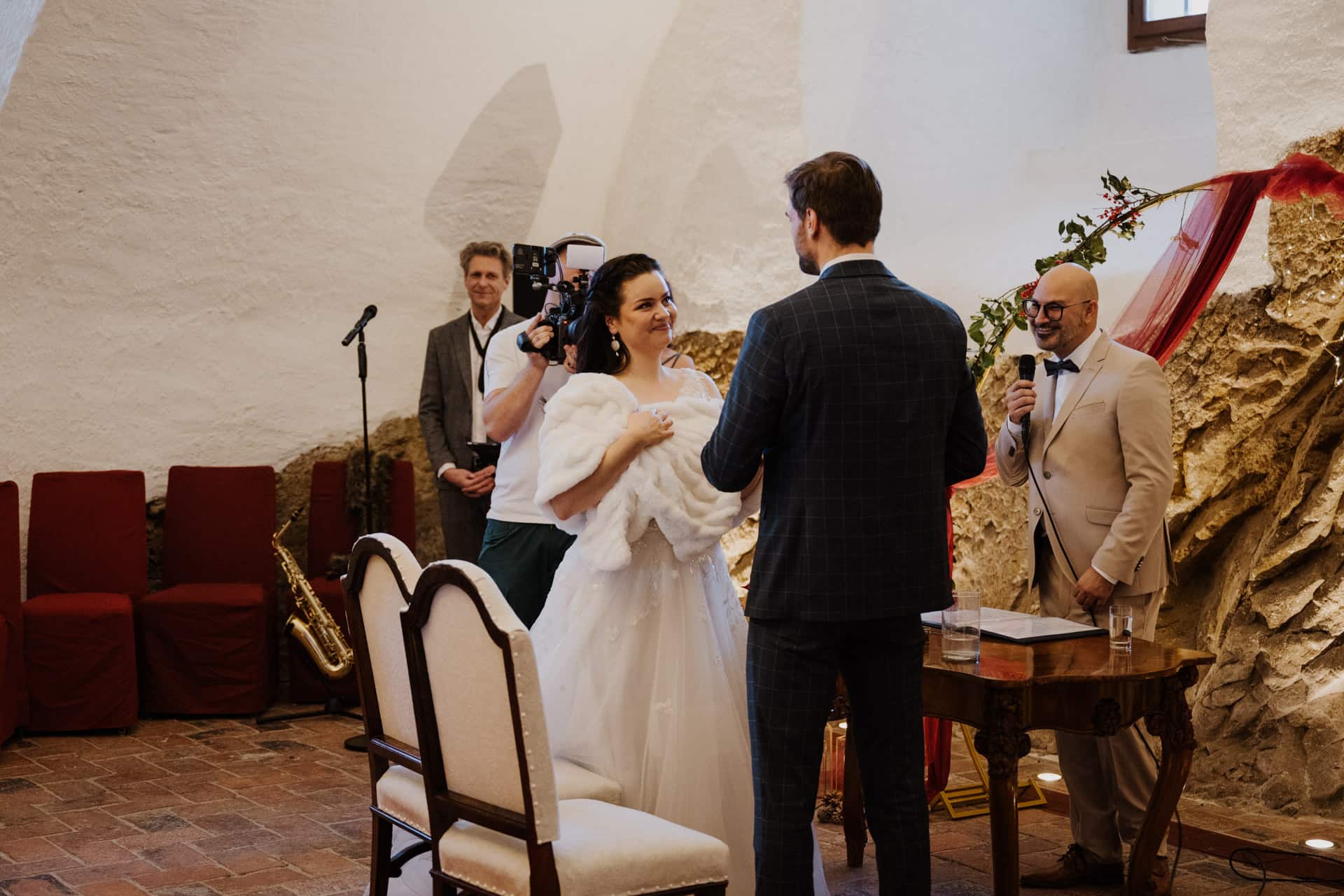Eine Braut und ein Bräutigam tauschen ihr Eheversprechen in einem rustikalen Innenraum aus, während ein lächelnder Beamter an einem kleinen Tisch sitzt und ein Kameramann den Moment aufnimmt. Der Raum hat Steinwände, rote Stühle und ein Saxophon steht in der Nähe.