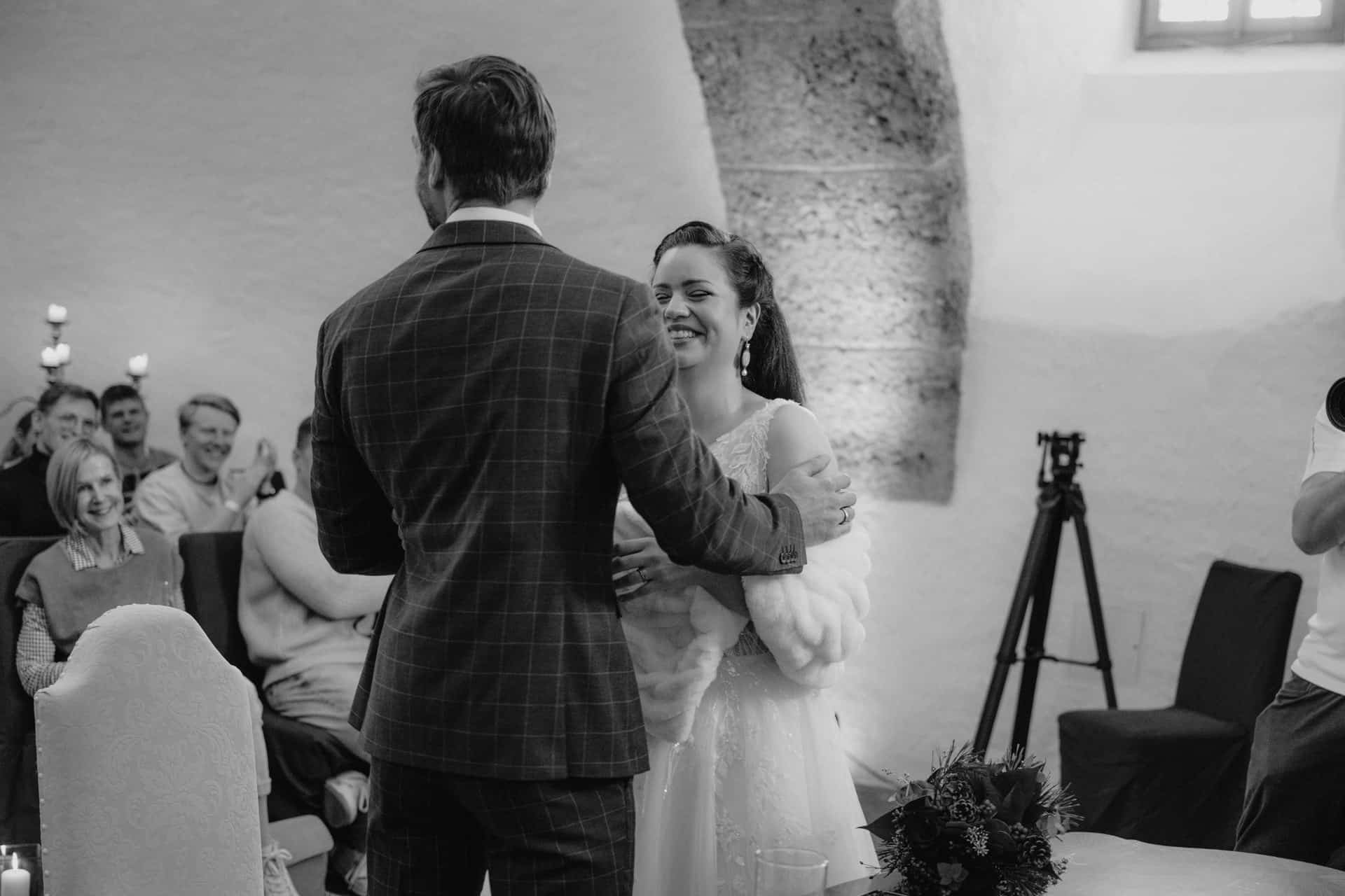 Braut und Bräutigam stehen lächelnd und umarmt während ihrer Hochzeitszeremonie, während die Gäste im Hintergrund in einem warm beleuchteten Raum zusehen und applaudieren.