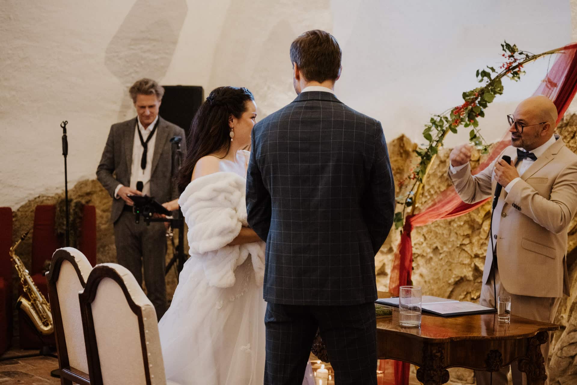 Eine Braut und ein Bräutigam stehen sich während einer Hochzeitszeremonie gegenüber, während ein Geistlicher neben ihnen spricht. Ein Musiker mit einem Saxophon steht im Hintergrund. Der Schauplatz ist ein Innenraum mit rustikalen, strukturierten Wänden.