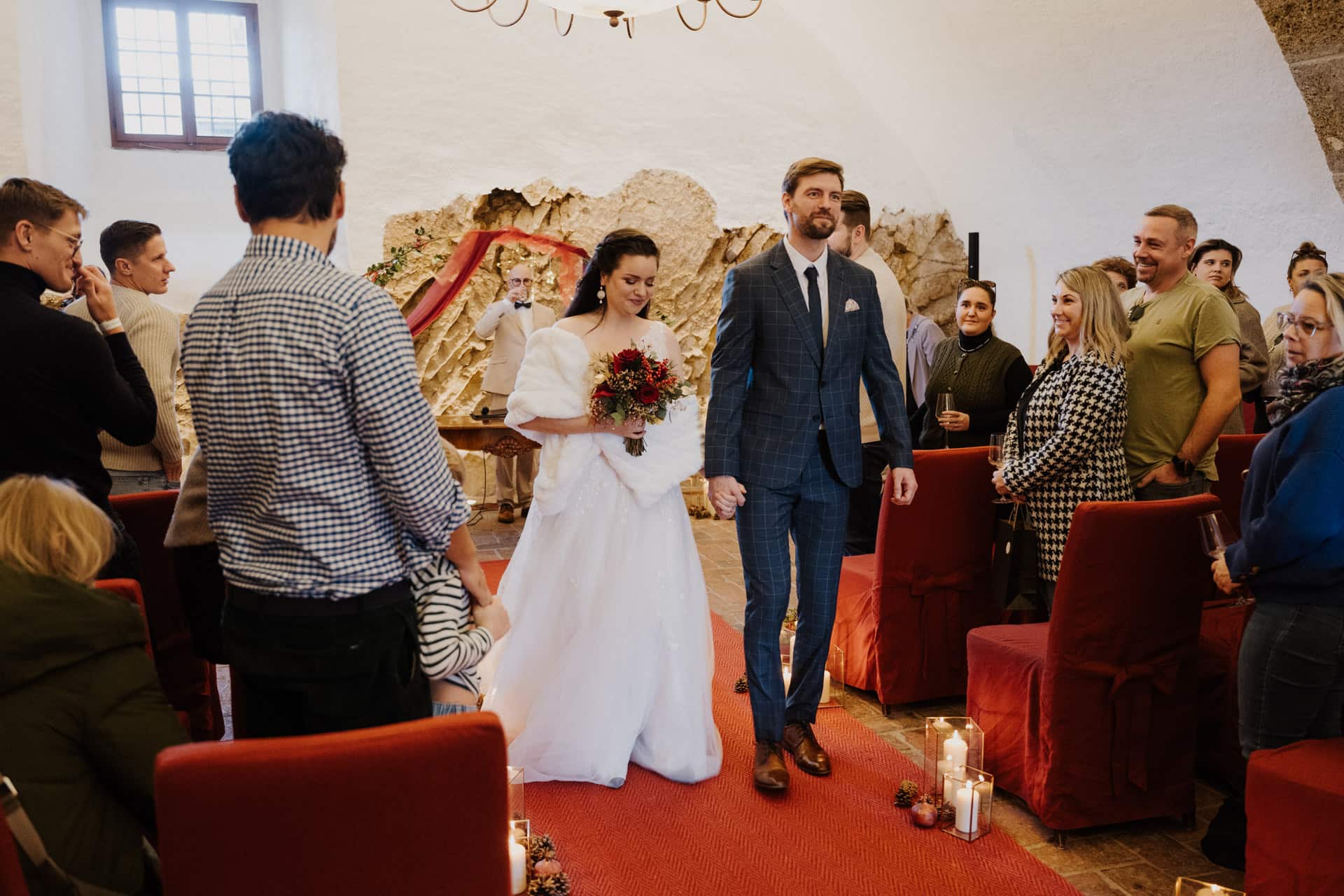 Eine Braut in einem weißen Kleid und ein Bräutigam in einem blauen Anzug schreiten händchenhaltend und lächelnd zum Altar, umgeben von sitzenden Gästen in einer warm beleuchteten Halle mit roten Akzenten und Steinwänden.