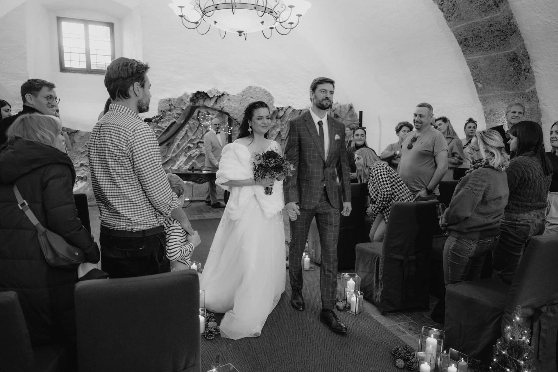 Braut und Bräutigam schreiten Hand in Hand in einem kerzenbeleuchteten Raum zum Altar und lächeln, während die sitzenden Gäste zusehen. Der Raum ist gemütlich mit gewölbten Decken und Steinwänden, die eine intime Hochzeitsatmosphäre schaffen.