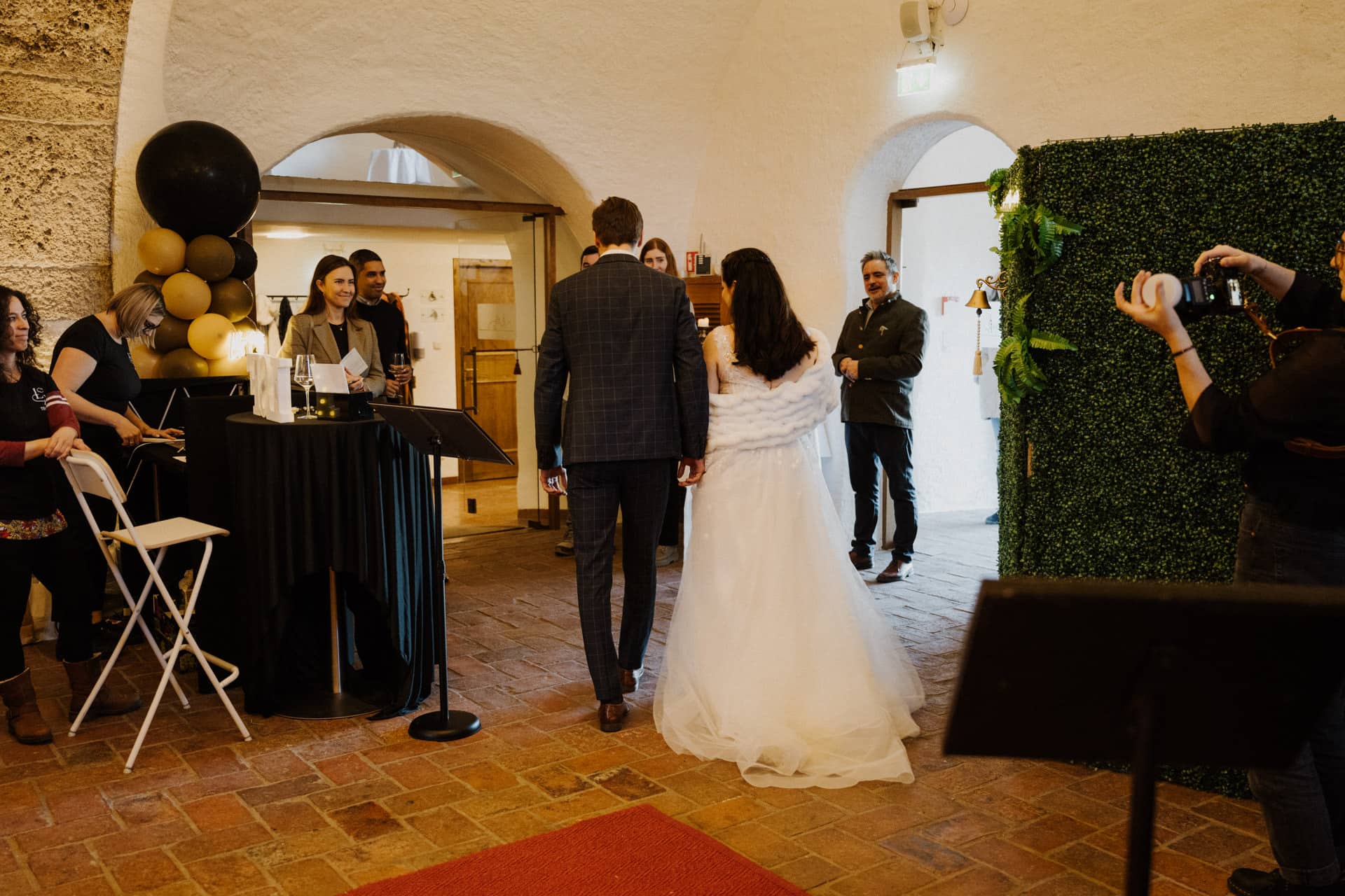 Eine Braut und ein Bräutigam betreten einen warm beleuchteten Veranstaltungsort mit weißen Steinwänden und werden von lächelnden Gästen begrüßt. Die Braut trägt ein weißes Kleid und ein Kopftuch, der Bräutigam einen karierten Anzug. Menschen stehen in der Nähe, Luftballons und eine begrünte Wand schmücken den Raum.