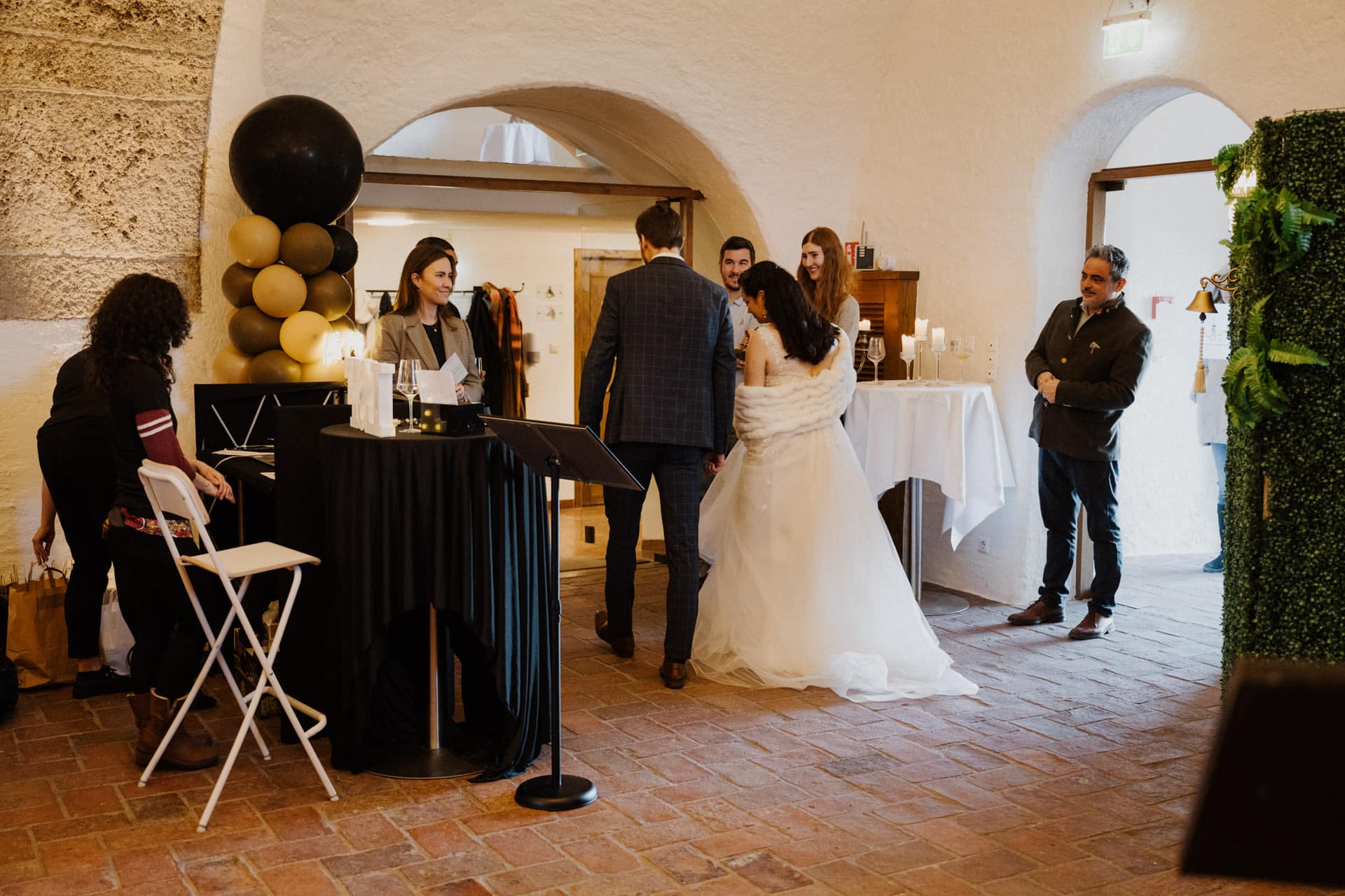 Eine Braut und ein Bräutigam stehen zusammen in einem warm beleuchteten Raum mit Steinwänden und unterhalten sich mit den Gästen einer mit schwarz-goldenen Ballons und Kerzen geschmückten Hochzeitsfeier. Links sitzt eine Frau an einer Tastatur.