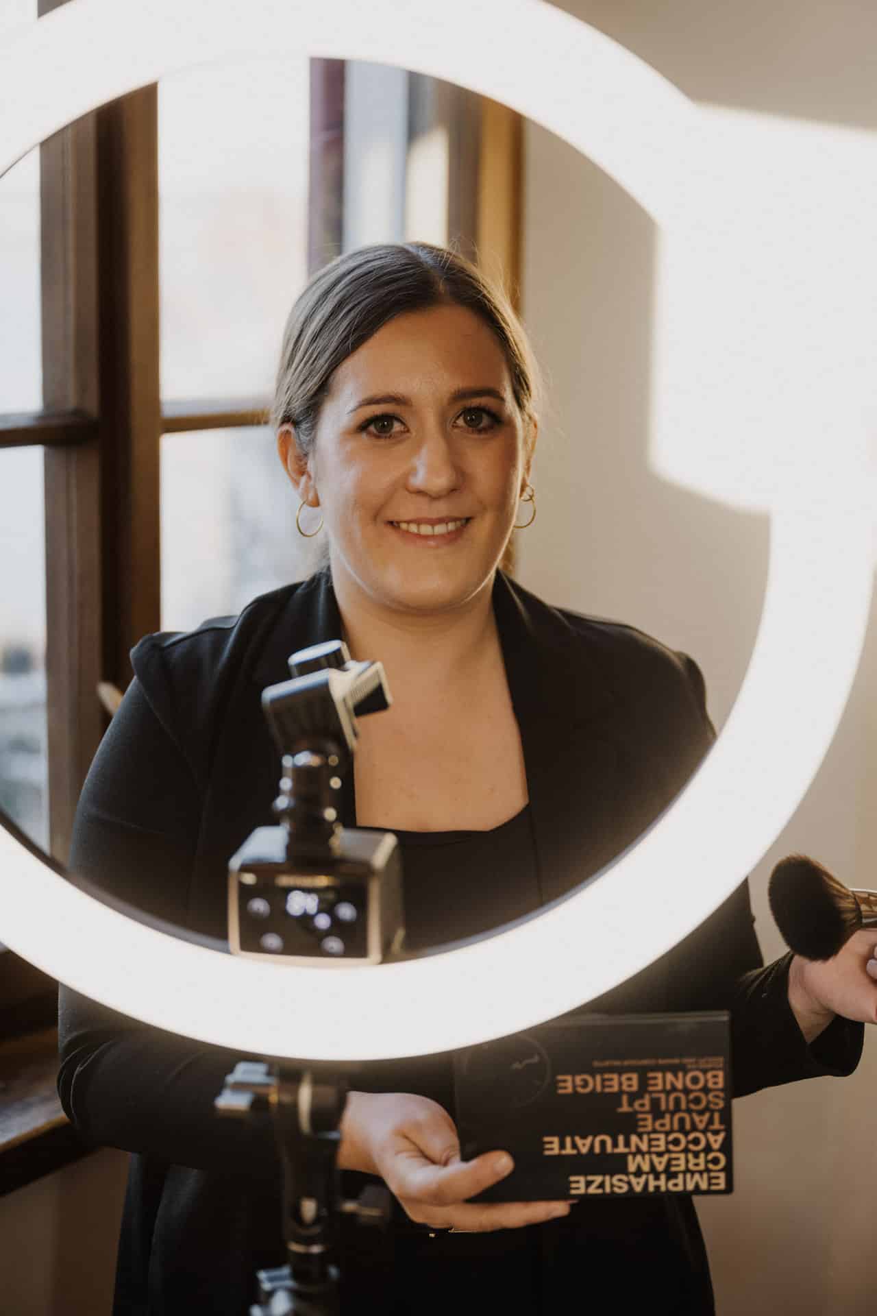 Eine Frau steht in einem kreisförmigen Lichtring und hält einen Schminkpinsel und ein Buch in der Hand. Sie lächelt in die Kamera, trägt einen schwarzen Blazer und goldene Ohrringe, während im Hintergrund eine Kamera und ein Fenster zu sehen sind.