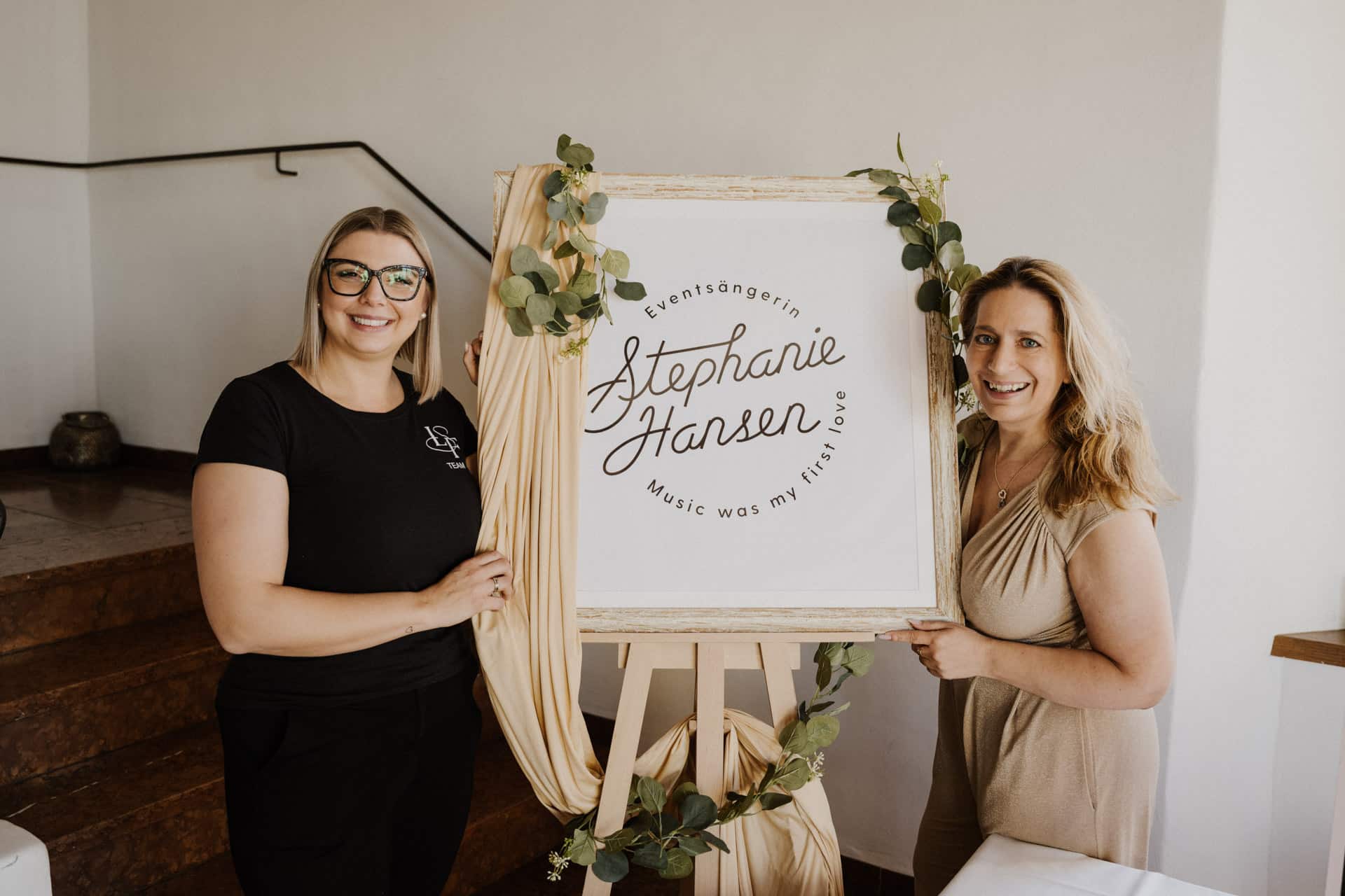 Zwei lächelnde Frauen stehen zu beiden Seiten eines geschmückten Schildes mit der Aufschrift "Event Sängerin Stephanie Hansen, Music was my first love" in einem Innenraum in der Nähe einer Treppe, die mit Grünpflanzen und drapierten Stoffen geschmückt ist.