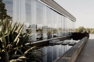 Ein modernes Gebäude von DasSee - Event Exclusive mit großen Glasfenstern, durchsichtigen weißen Vorhängen und einem schmalen reflektierenden Pool mit quadratischen Trittsteinen, umgeben von üppigen Pflanzen und Bäumen unter einem klaren Himmel.