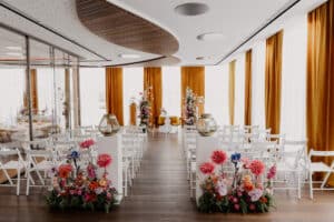 Eine moderne DasSee - Event Exclusive Hochzeitszeremonie mit weißen Stühlen in Reihen, farbenfrohen Blumenarrangements entlang des Ganges, großen goldenen Vasen und senfgelben Vorhängen, die helle Fenster umrahmen.
