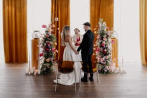 Eine Braut und ein Bräutigam stehen händchenhaltend vor einem Zeremonienmeister während einer DasSee - Event Exclusive Hochzeitszeremonie, umgeben von farbenfrohen Blumenarrangements und Kerzen, mit goldenen Vorhängen im Hintergrund.