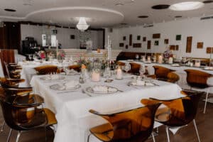 Eleganter Essbereich mit weißen Tischdecken, Blumenschmuck, Kerzen und bernsteinfarbenen Stühlen, die um gedeckte Tische herum angeordnet sind - perfekt für eine DasSee - Event Exclusive-Veranstaltung in einem modernen, sanft beleuchteten Restaurantinterieur.