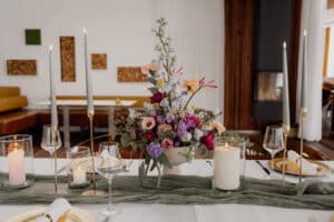 Elegant DasSee - Event Exklusiver Esstisch mit weißen Tellern, Gläsern und goldenen Utensilien, mit hohen Kerzen, einem grünen Tischläufer und einem farbenfrohen floralen Mittelstück in einem modernen, gemütlichen Raum.