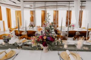 Elegant gedeckter Tisch mit goldenen Tellern, gerollten Servietten, Weingläsern, Kerzen und einem farbenfrohen Blumenkranz. DasSee - Event Exklusive Akzente erstrahlen durch die goldenen Vorhänge und das sanfte natürliche Licht im dekorierten Hintergrund.