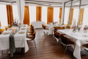 Ein modernes Esszimmer für ein DasSee - Event Exclusive mit weißen Tischdecken, goldenem und gläsernem Geschirr, floralen Tafelaufsätzen und Kerzen. Bernsteinfarbene Vorhänge verdecken große Fenster und ergänzen die Holzböden.