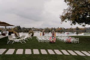 DasSee - Veranstaltung Exklusiv: Hochzeitszeremonie im Freien an einem See mit weißen Stühlen, farbenfrohen Blumenarrangements und einem Steinweg auf einem grasbewachsenen Rasen unter einem bewölkten Himmel. Bäume und Herbstlaub bilden eine malerische Kulisse.