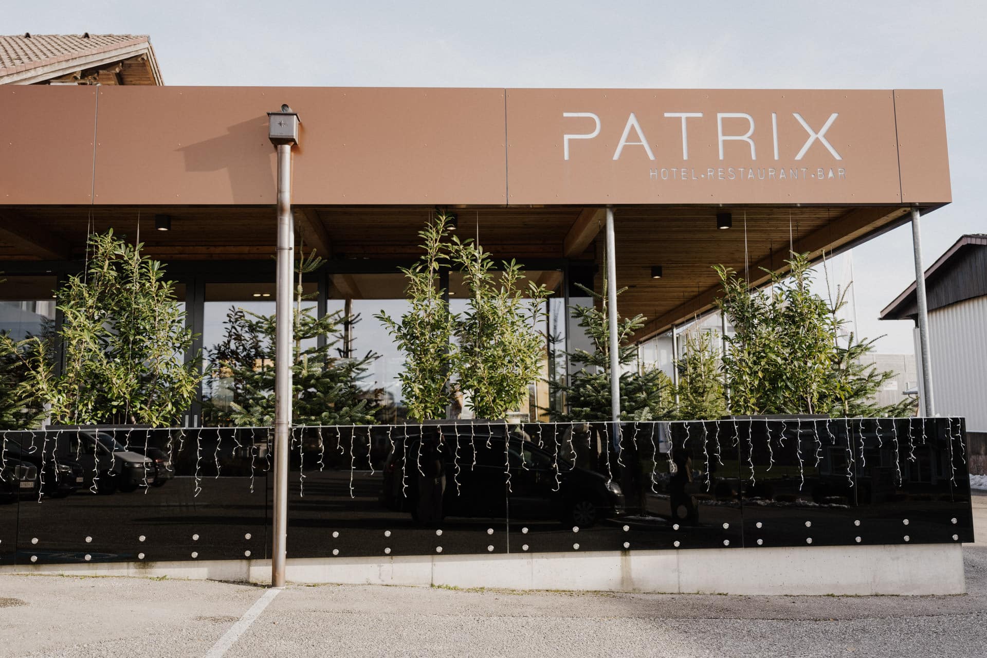 Ein modernes Gebäude mit einem braunen Schild mit der Aufschrift "PATRIX HOTEL.RESTAURANT.BAR". Vor dem Eingang stehen kleine Bäume und Lichterketten entlang einer Glaswand. Der Himmel ist klar und die Gegend ist gut beleuchtet.