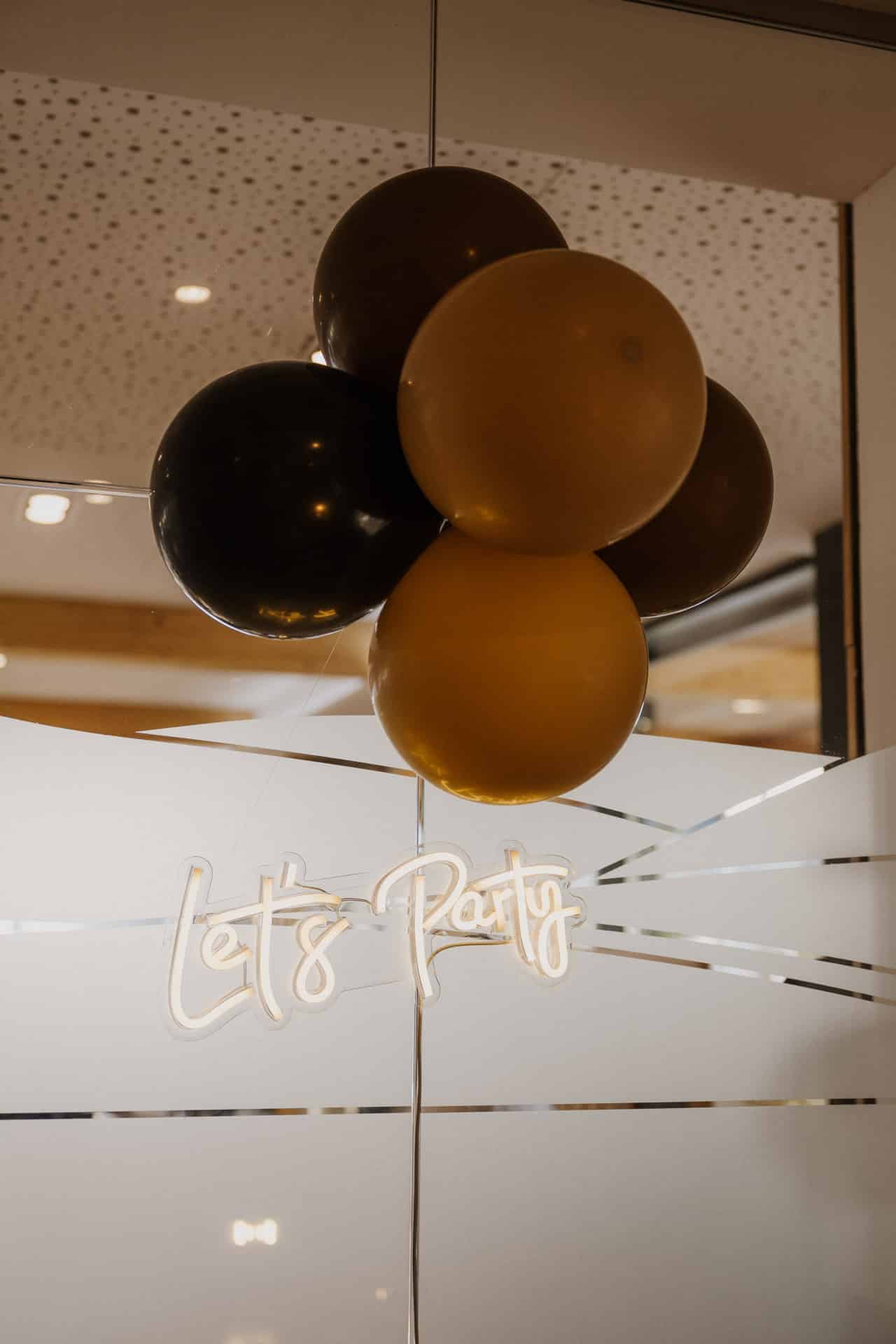 Ein Bündel schwarzer und goldener Luftballons hängt über einem leuchtenden Neonschild mit der Aufschrift "Let's Party", das an einer Glaswand mit mattierten Streifen angebracht ist.