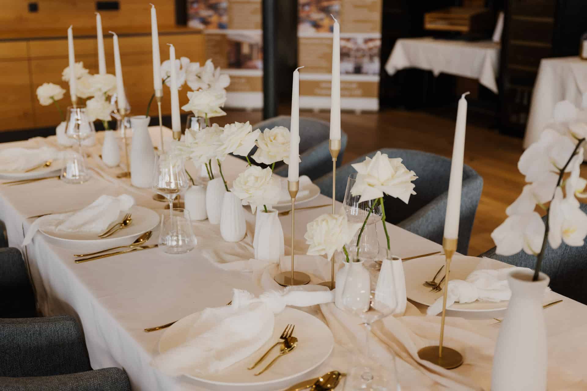 Elegant gedeckter Tisch mit weißen Blumen in Vasen, hohen weißen Kerzen, weißen Servietten und goldenem Besteck auf einem weißen Tischtuch, umgeben von blauen Stühlen in einem warm beleuchteten Raum.