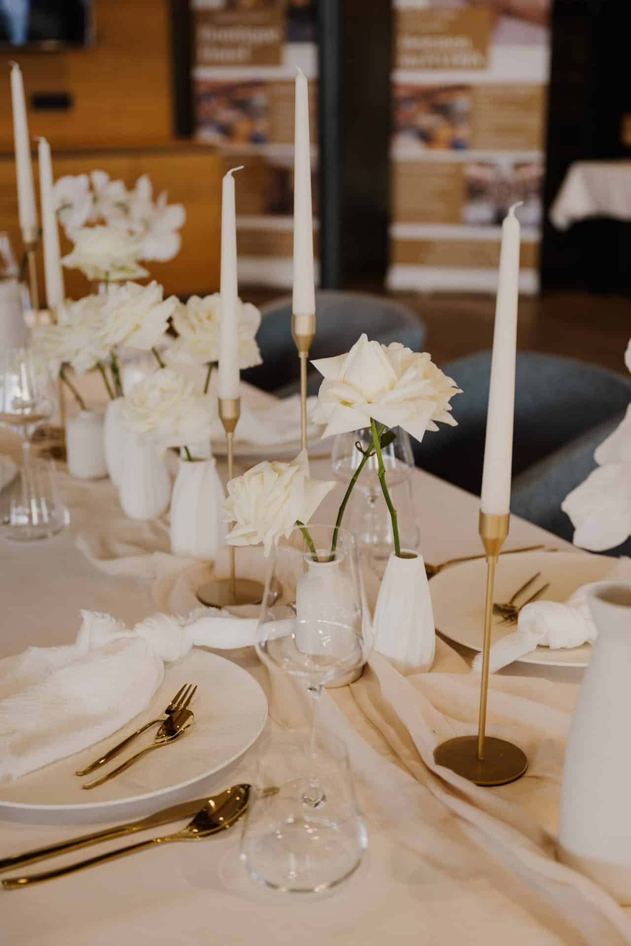 Ein elegant gedeckter Tisch mit weißen Tellern, goldenem Besteck, weißen Servietten, hohen weißen Kerzen in goldenen Haltern und weißen Blumen in kleinen Vasen sorgt für ein raffiniertes und minimalistisches Dekor.
