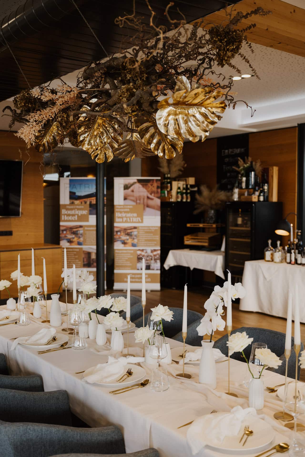 Ein stilvoll gedeckter Essbereich für eine Veranstaltung mit einem langen Tisch mit weißen Blumen, Kerzen und eleganten Gedecken. Über dem Tisch hängt ein dekoratives Arrangement mit goldenen Blättern und getrockneten Zweigen.