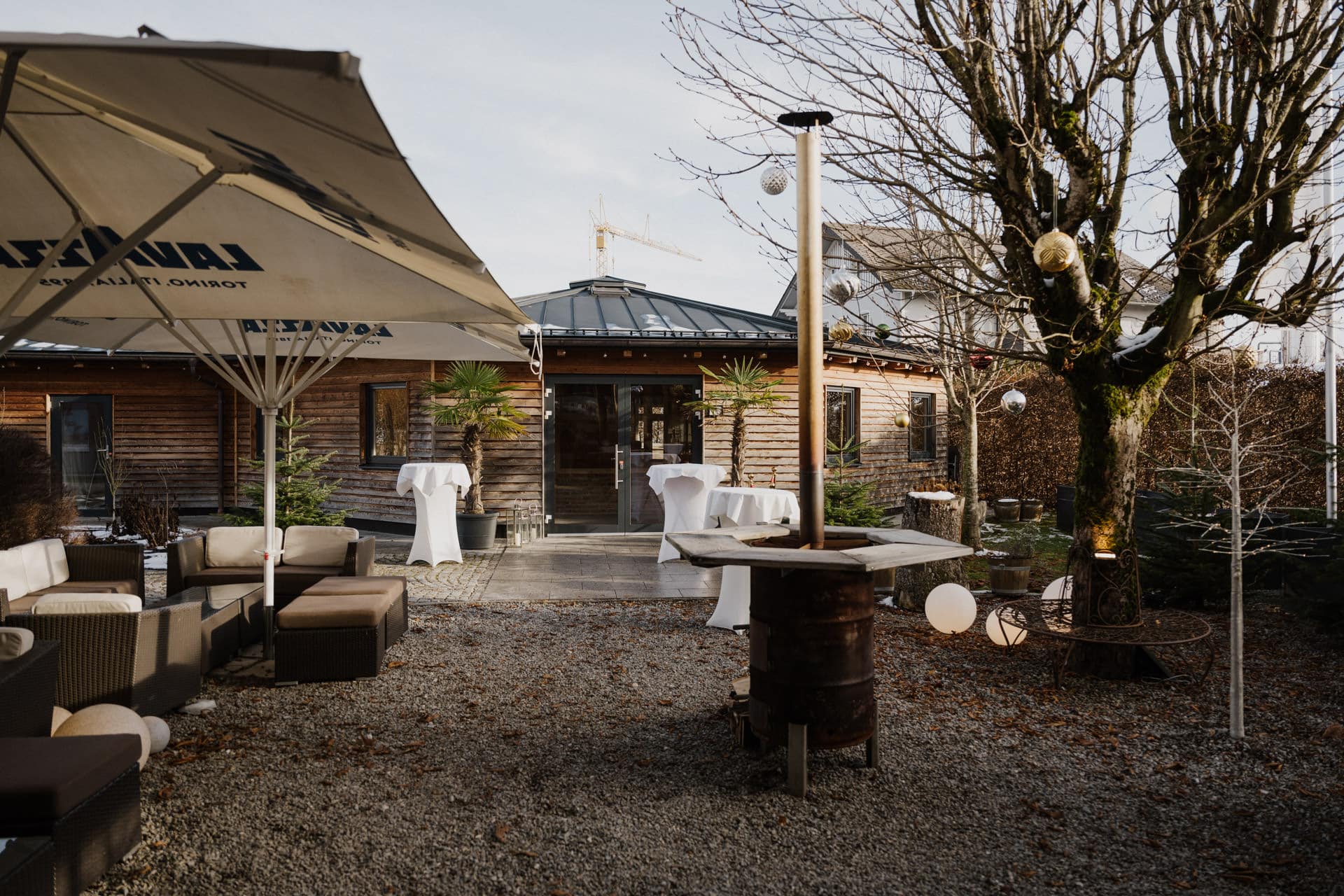 Terrasse im Freien mit Kiesboden, runden, mit weißem Tuch bedeckten Cocktailtischen, einem Holzgebäude im Hintergrund, einem blattlosen Baum, einem großen Sonnenschirm und dekorativen Lichtern in den Zweigen.