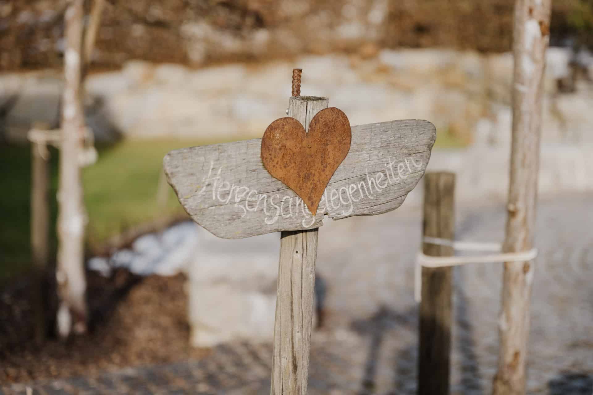 Ein hölzernes Schild mit einem verrosteten Metallherz und der handschriftlichen Aufschrift "Herzensbegegnungen" steht an einem sonnigen Tag im Freien, mit verschwommenem Grün und Stein im Hintergrund.