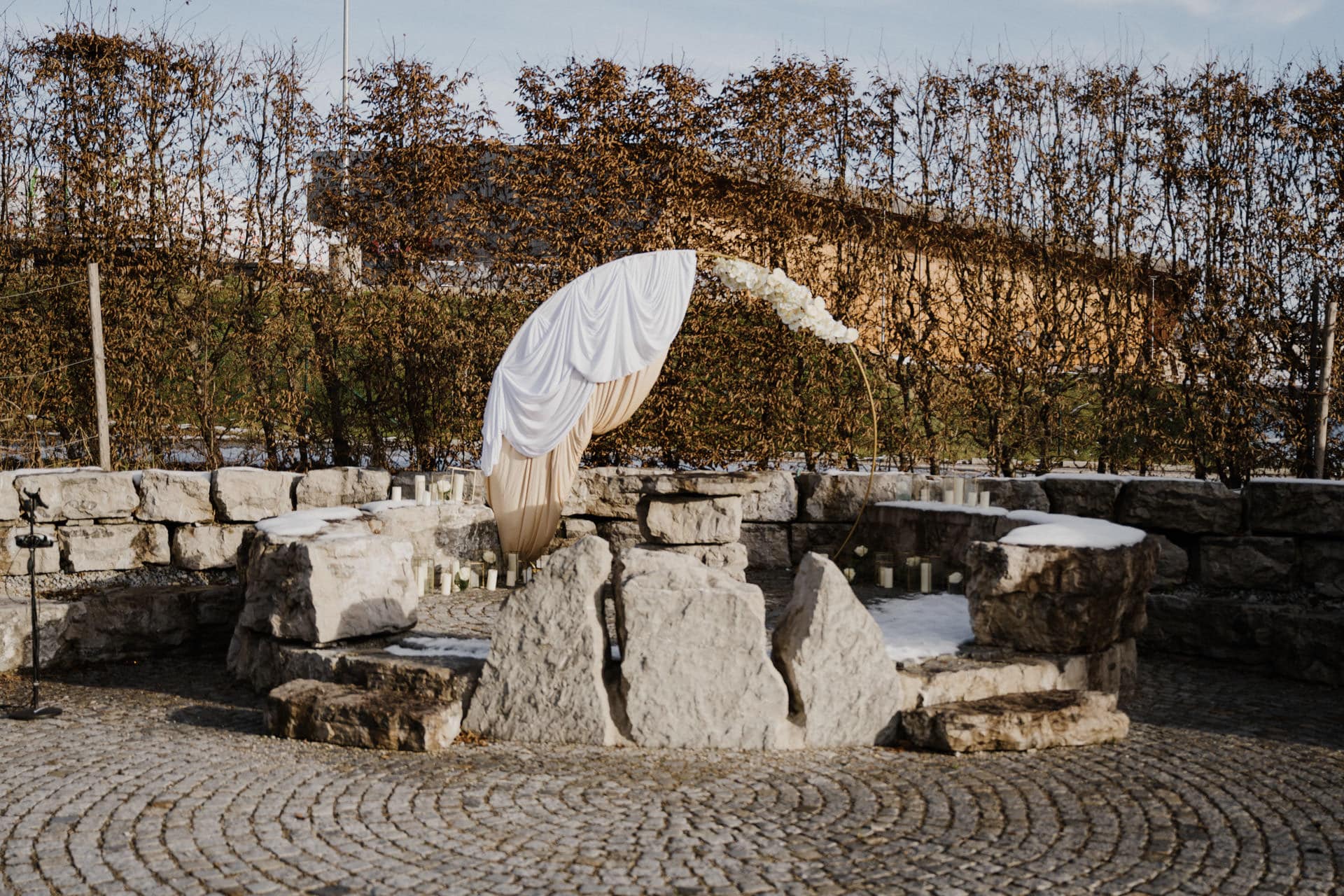 Ein Hochzeitsaltar im Freien ist mit weißem und beigem Stoff und weißen Blumen auf einem runden Metallbogen drapiert, der zwischen großen Steinplatten mit Kerzen steht, umgeben von Steinmauern und kahlen Bäumen im Winter.