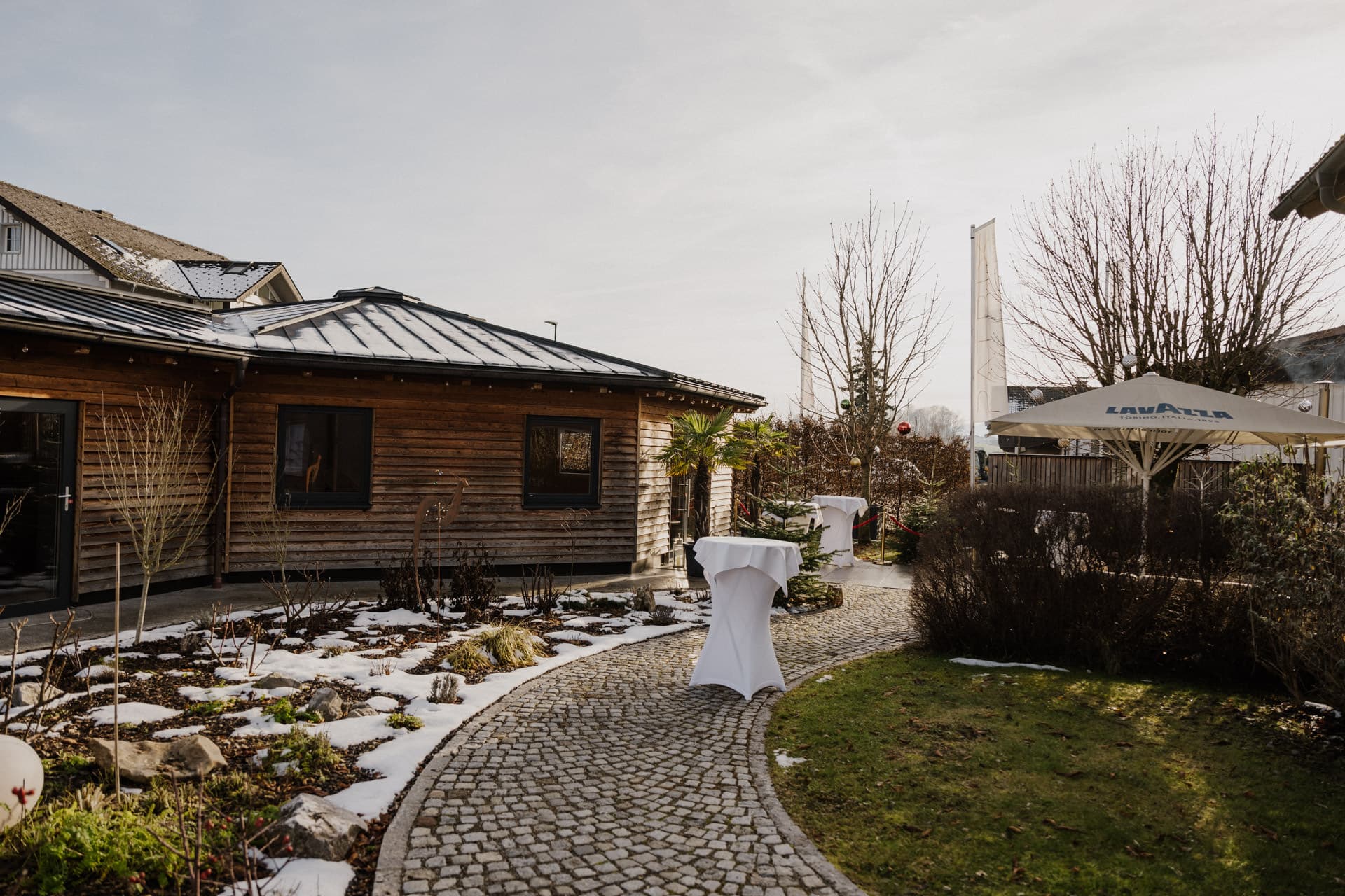 Ein Steinweg schlängelt sich durch einen Garten mit Schneeflecken und führt zu einem Holzgebäude. Überdachte Cocktailtische und ein großer Sonnenschirm im Freien deuten auf eine Versammlung oder eine Veranstaltung hin. Im Hintergrund stehen blattlose Bäume.