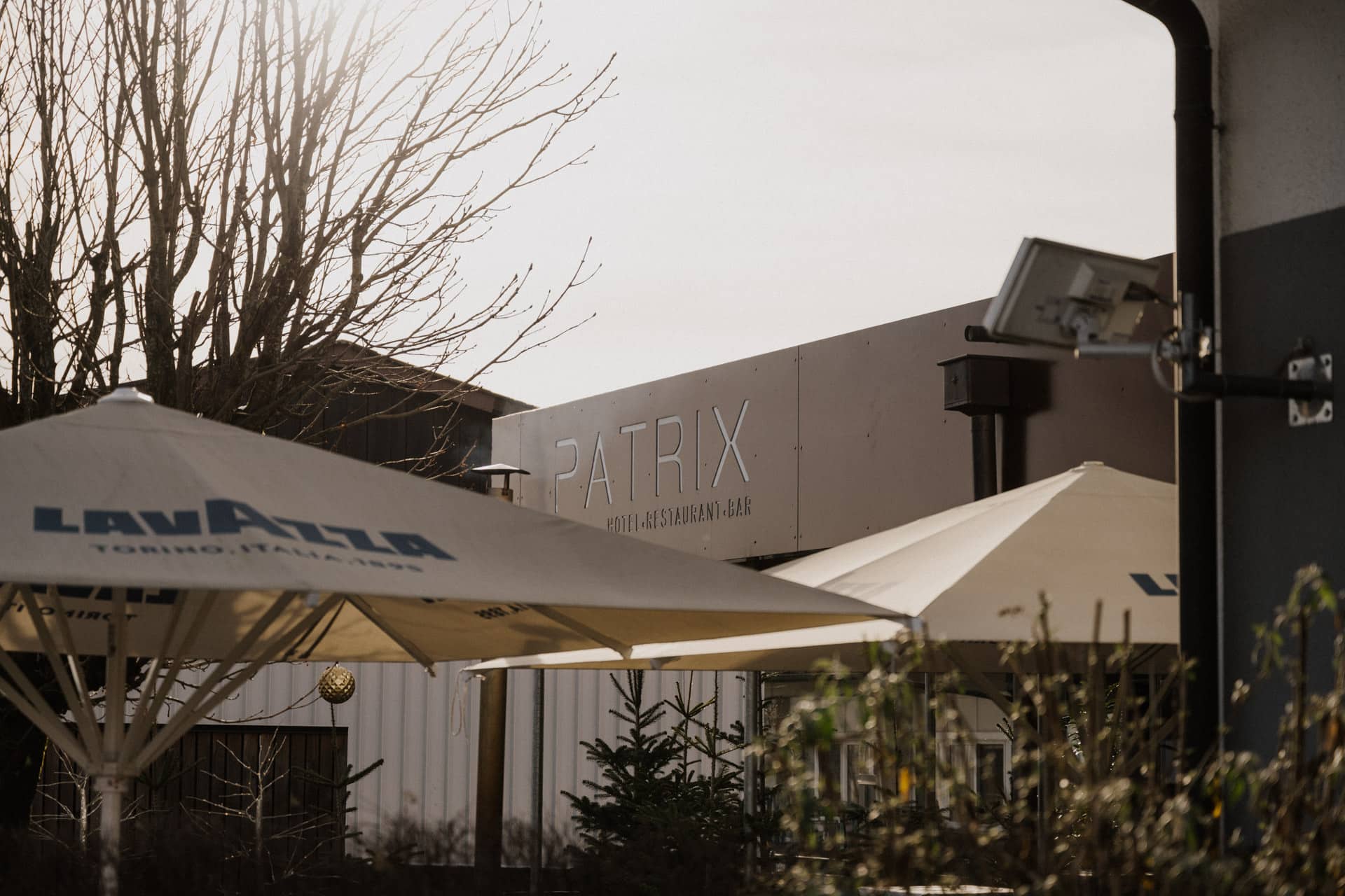 Außenansicht eines Restaurants namens "Patrix" mit großen beigen Sonnenschirmen mit der Aufschrift "Lavazza" und kahlen Baumzweigen, die sich gegen den bewölkten Himmel abheben.