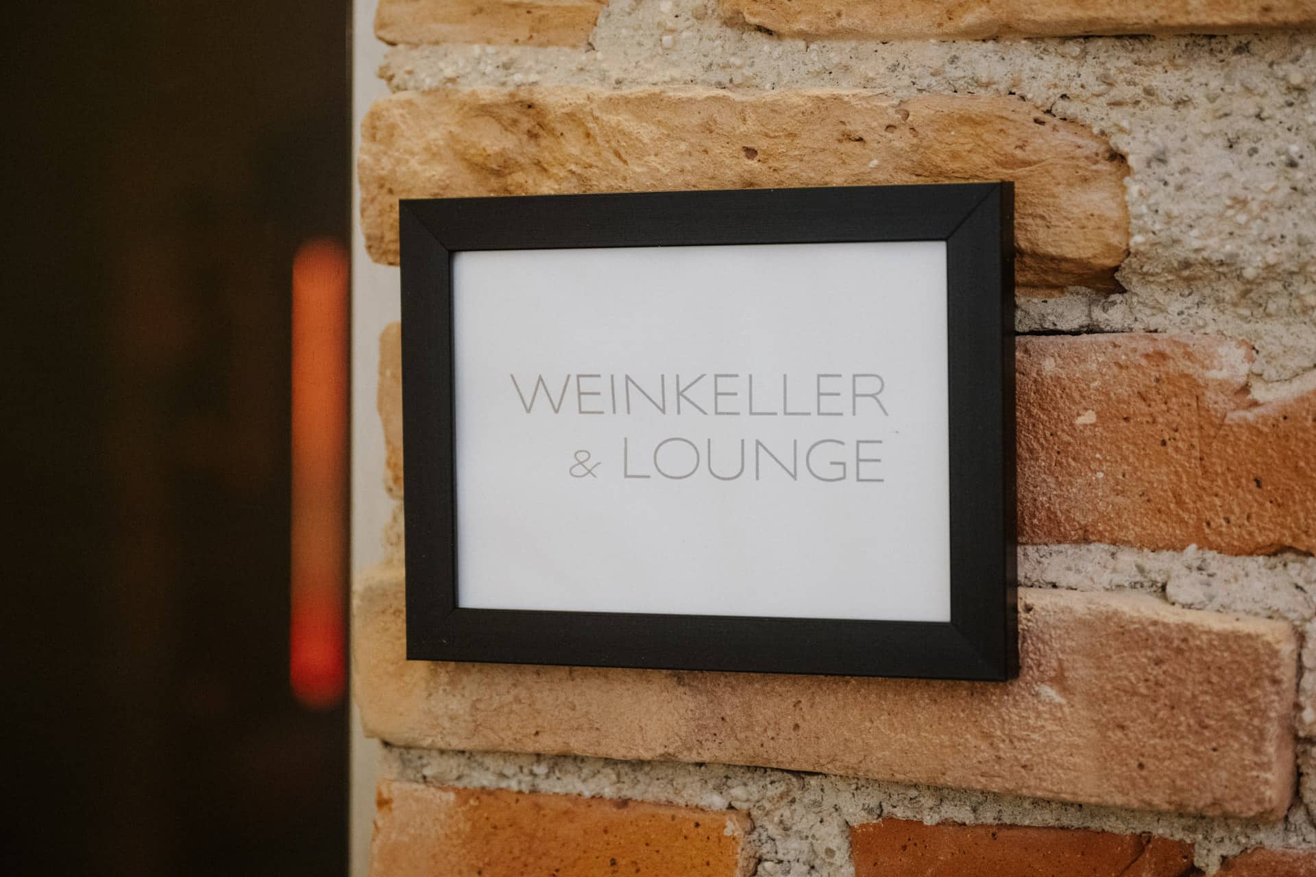 Auf einem gerahmten Schild an einer Backsteinwand steht in hellgrauen Großbuchstaben "WEINKELLER & LOUNGE".