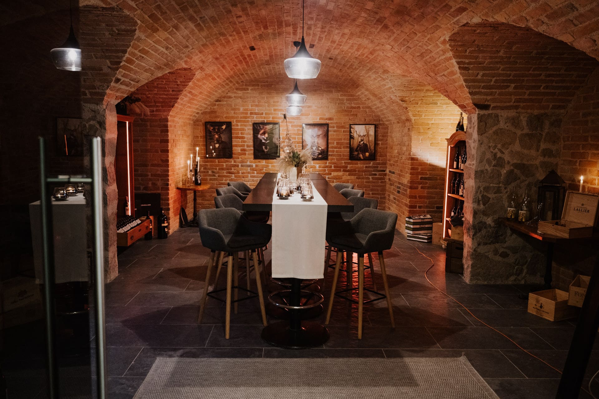Ein gemütlicher Weinkeller mit Backsteinwänden und gewölbter Decke, an dem ein langer Tisch zum Essen gedeckt ist, umgeben von gepolsterten Hockern, warmer Beleuchtung, Kerzen und gerahmter Kunst an den Wänden.