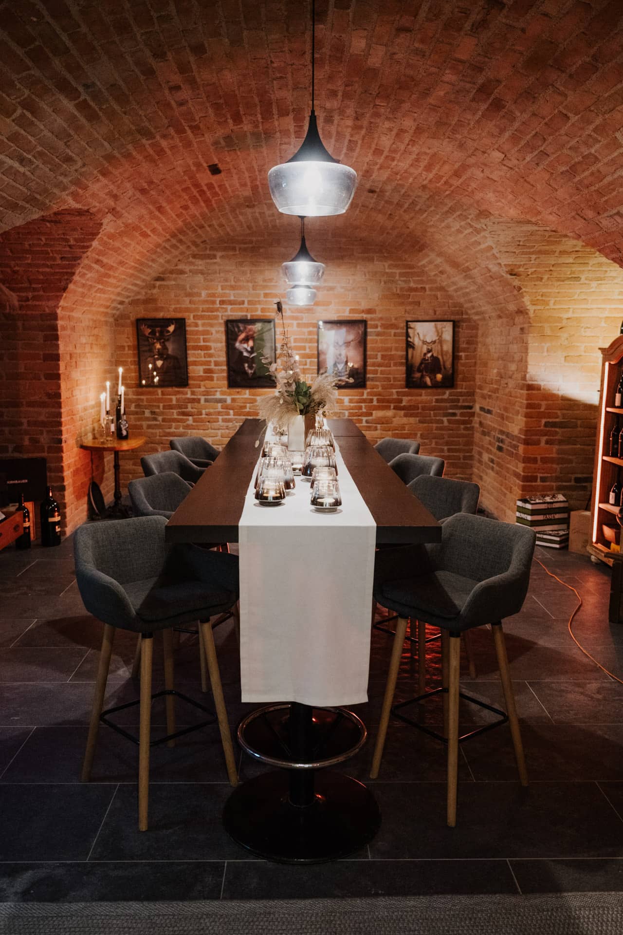 In einem gemütlichen Raum mit Backsteingewölbe, modernen grauen Stühlen, Pendelleuchten, Kerzen und gerahmter Kunst an den Wänden ist ein langer Esstisch mit weißem Läufer für die Mahlzeit gedeckt.