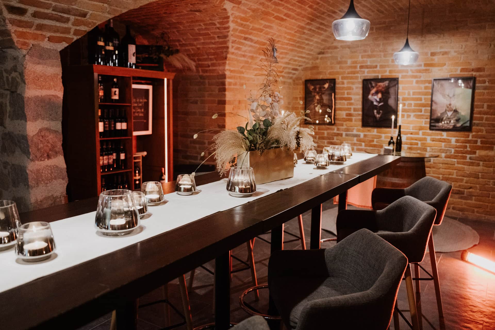 Ein stilvoller Weinkeller mit Backsteinwänden, einem langen Tisch mit Kerzen und einem Tafelaufsatz, gepolsterten Stühlen, Regalen mit Weinflaschen und modernen Pendelleuchten, die eine gemütliche Atmosphäre schaffen.