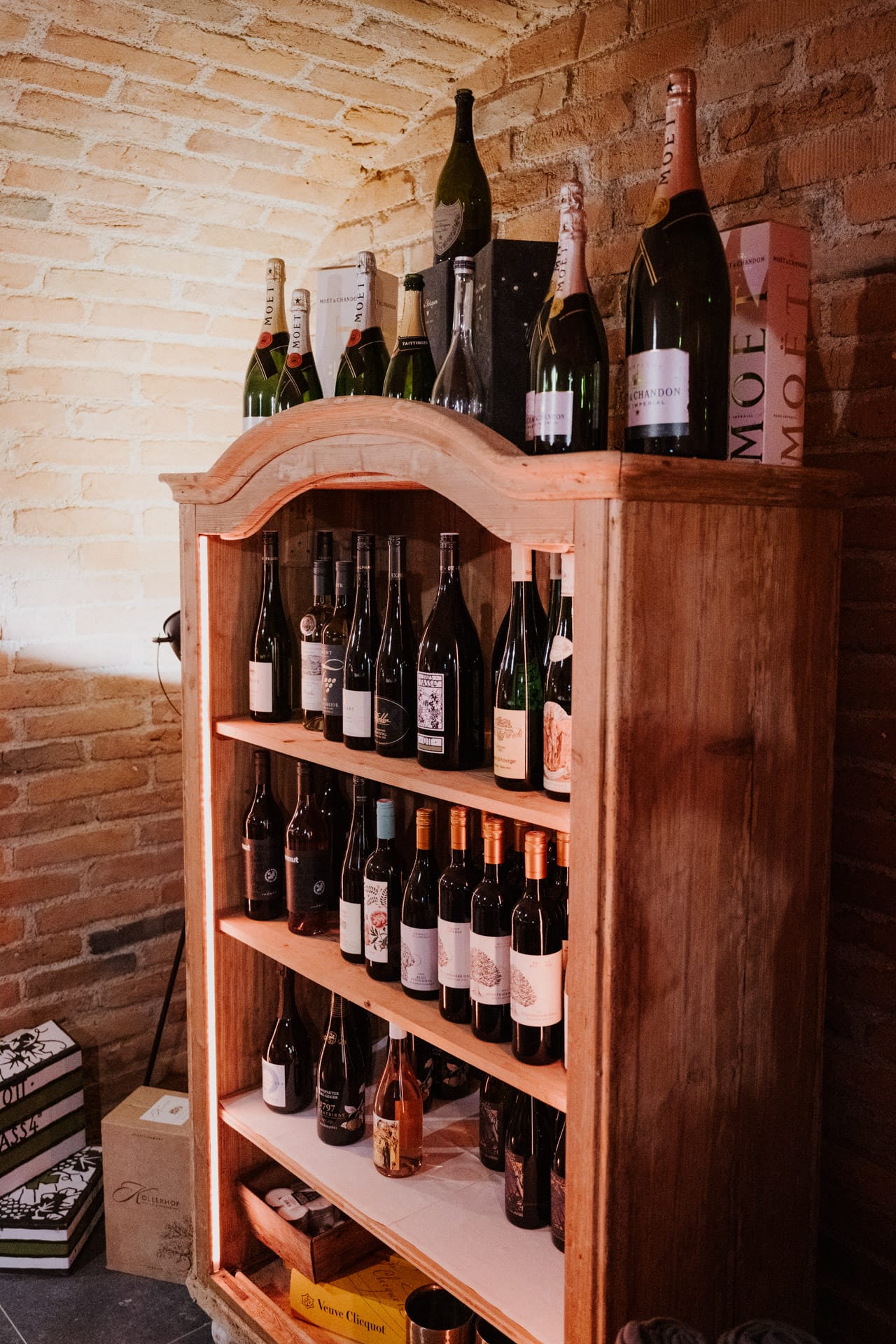 Holzschrank, gefüllt mit verschiedenen Wein- und Sektflaschen, sowohl oben als auch auf den Regalen, der in einem schwach beleuchteten Raum an einer Backsteinwand steht. Auf dem Boden neben dem Schrank sind weitere Kisten zu sehen.