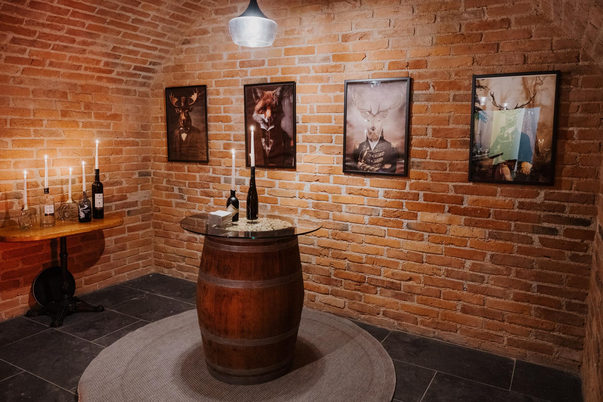 Ein gemütlicher, gemauerter Weinkeller mit einem runden Fasstisch, Weinflaschen und Kerzen. An der Wand hängen vier gerahmte Tierporträts, links daneben steht ein kleiner Holztisch mit Kerzen und Flaschen.