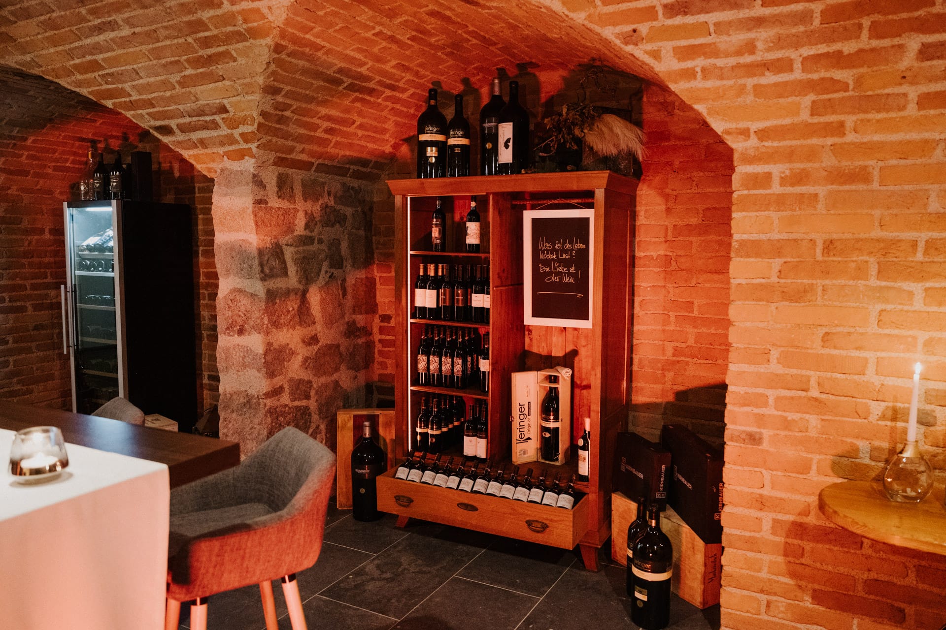 Ein gemütlicher Weinkeller mit Backsteinwänden, Holzregalen voller Weinflaschen, einem grauen Stuhl, einem kleinen Tisch und warmer Beleuchtung, die eine einladende Atmosphäre schafft.