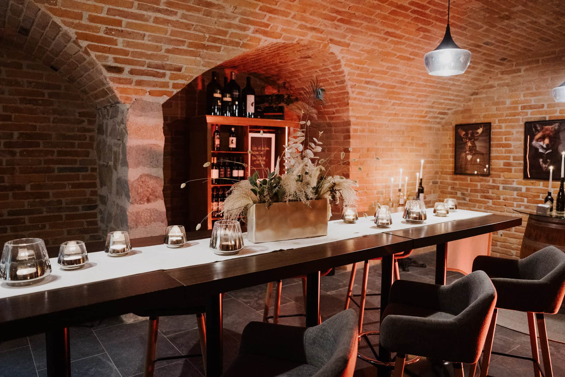 Ein gemütlicher Weinkeller mit Backsteinwänden, ein langer Tisch mit Kerzen und Blumenschmuck, umgeben von modernen grauen Stühlen. Im Hintergrund sind Weinflaschen in Regalen ausgestellt, die von warmen Pendelleuchten beleuchtet werden.