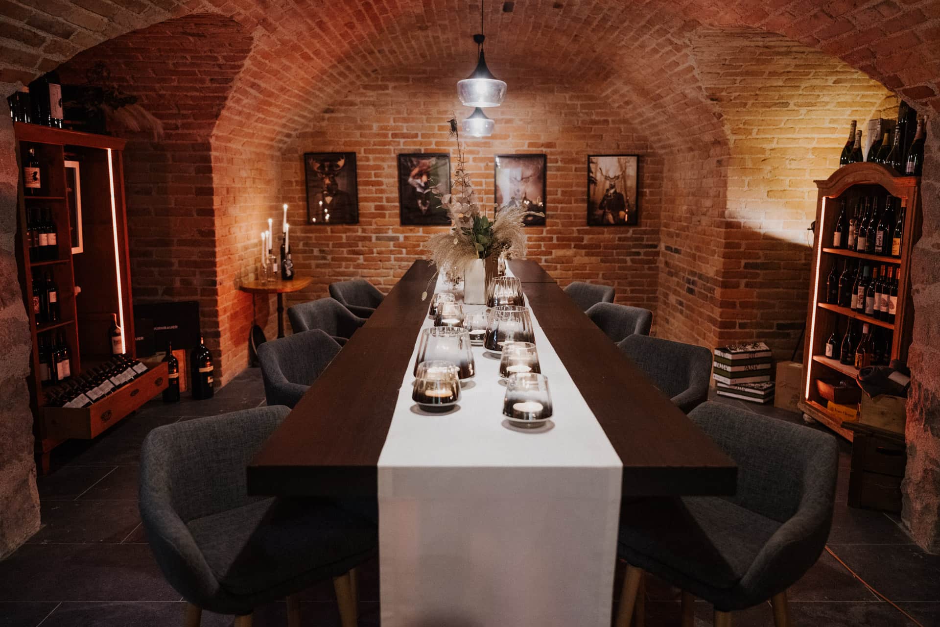 Ein gemütlicher Weinkeller mit Backsteinwänden und gewölbter Decke, an dem ein langer Holztisch mit Kerzen und Tafelaufsätzen gedeckt ist, umgeben von gepolsterten Stühlen und mit Weinflaschen gefüllten Regalen. Die warme Beleuchtung schafft eine einladende Atmosphäre.