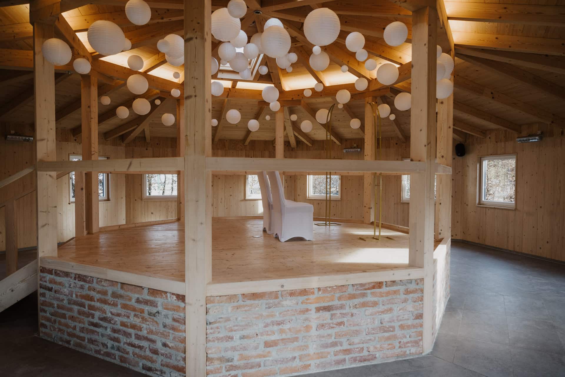 Ein geräumiger, runder Holzraum mit gemauertem Sockel, mit einem erhöhten Podest in der Mitte, weißen Papierlaternen, die von der Decke hängen, und einem einzelnen weißen Stuhl in der Mitte. Große Fenster lassen viel natürliches Licht herein.