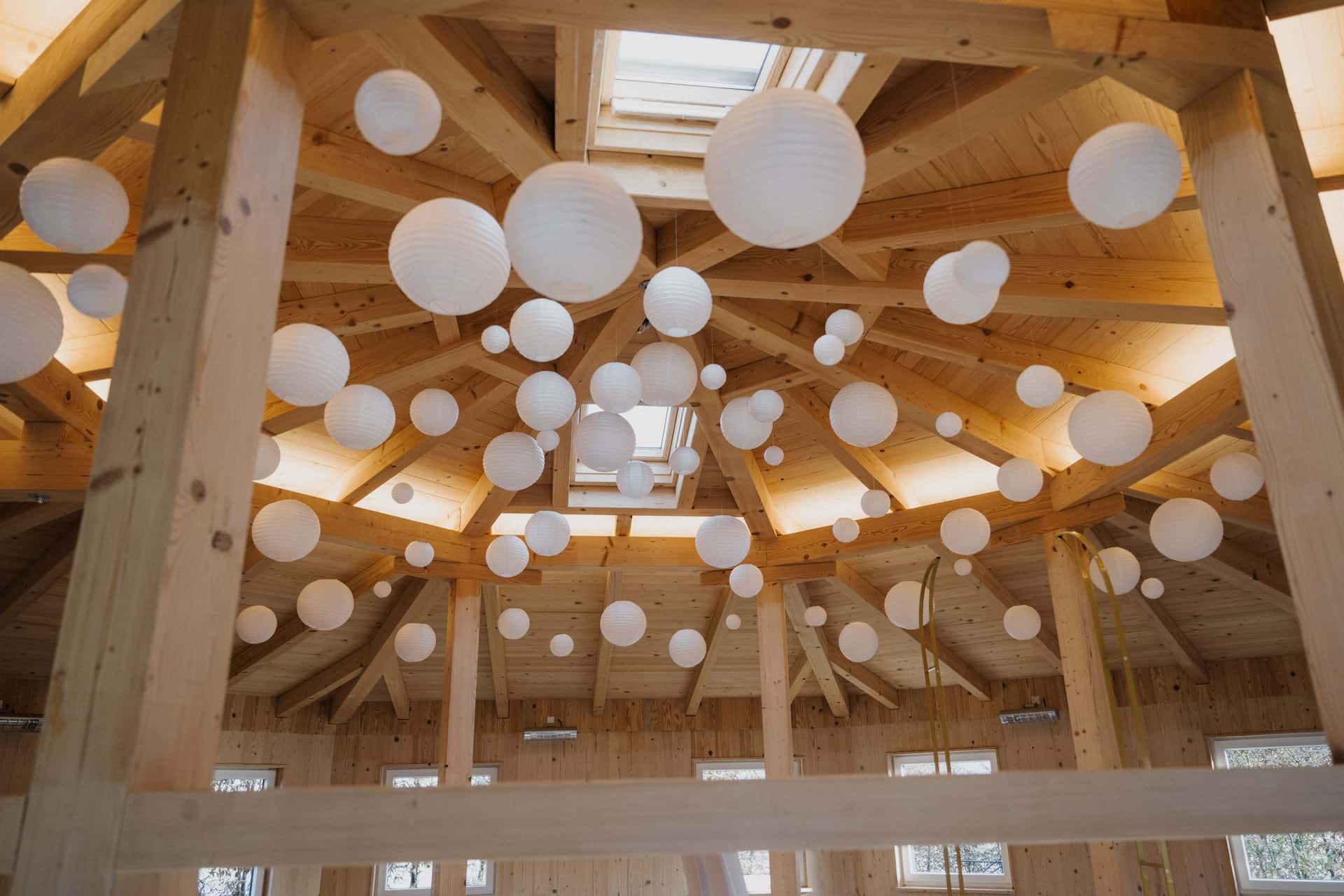 Eine Holzdecke mit sichtbaren Balken ist mit zahlreichen weißen Papierlaternen verziert, die in verschiedenen Höhen hängen. Sonnenlicht fällt durch ein Oberlicht ein und erhellt den gemütlichen, unfertigen Raum mit mehreren Fenstern.