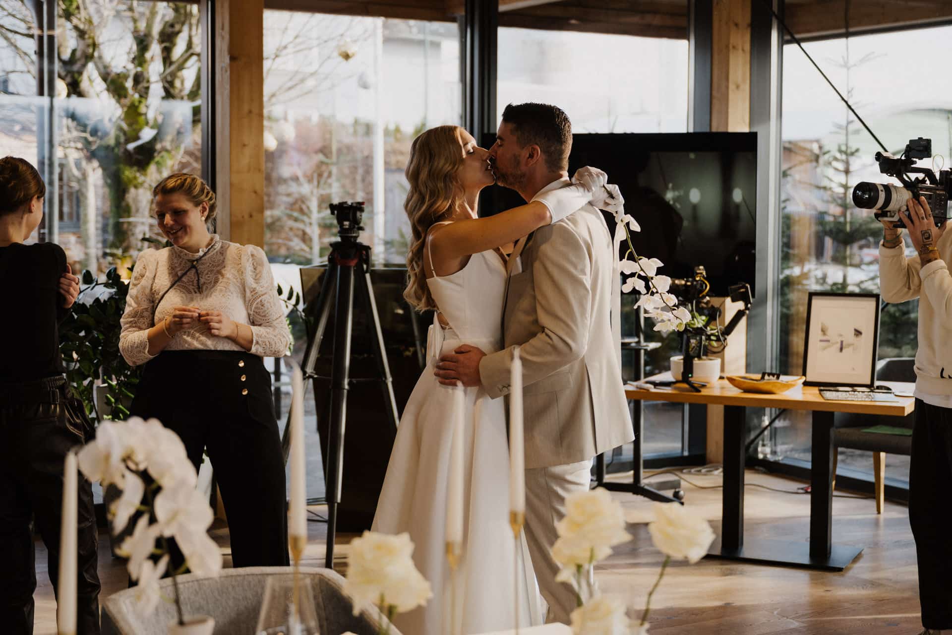 Braut und Bräutigam küssen sich auf der Tanzfläche einer Hochzeitsfeier, umgeben von Gästen, Kameras und Blumendekorationen in einem hellen, modernen Lokal.