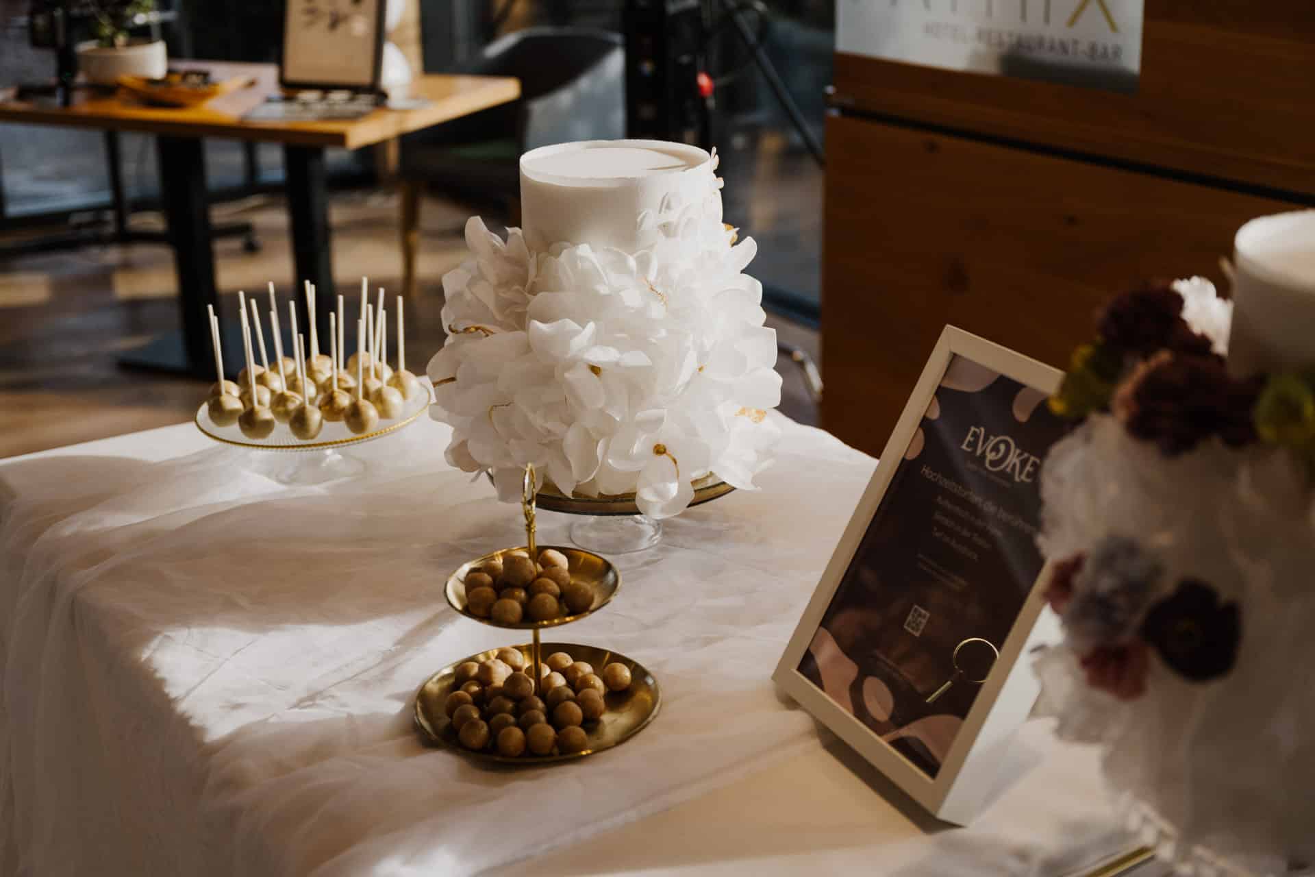 Eine weiße Torte, die mit großen weißen Blumen verziert ist, steht auf einem Tisch neben Cake Pops, goldenen Tabletts mit runden Süßigkeiten und einem gerahmten Schild, alles auf einem weißen Tischtuch.