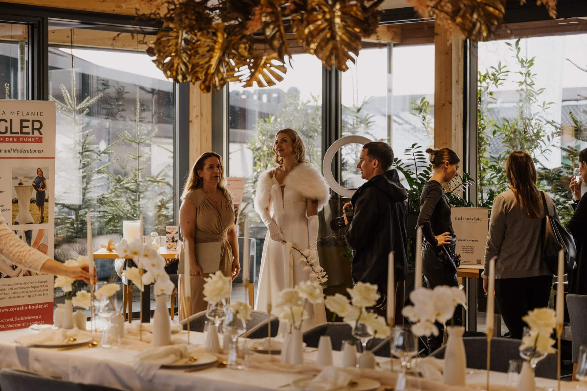 In einem modernen, sonnendurchfluteten Raum mit großen Fenstern und Holzbalken steht eine Gruppe von Menschen um einen mit weißen Blumen und Kerzen gedeckten Tisch herum und unterhält sich.