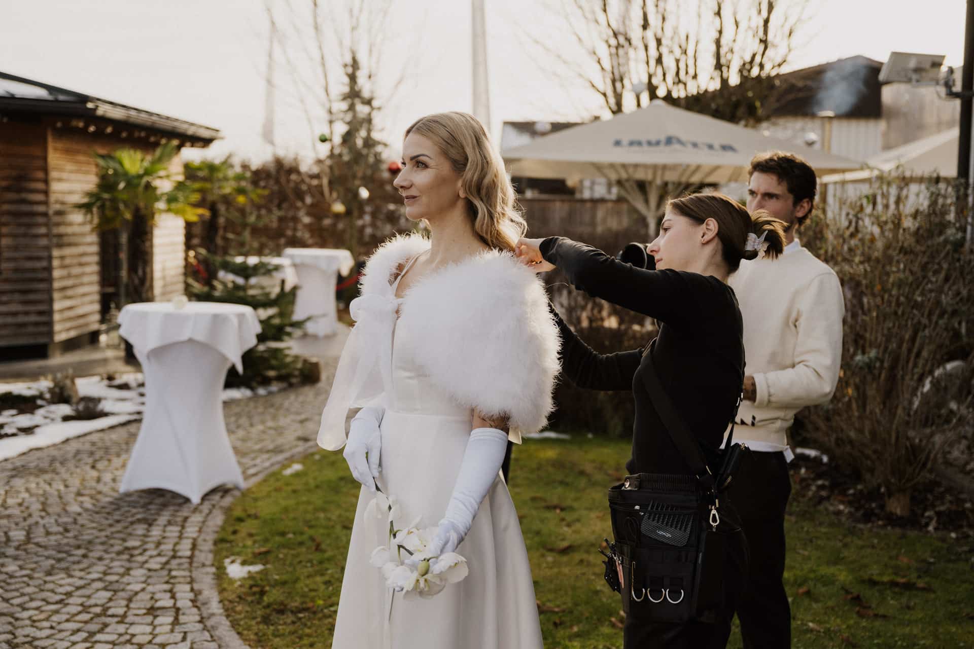Eine Braut im weißen Kleid und mit Handschuhen steht im Freien, während eine Stylistin ihren Schal zurechtrückt; ein Mann in legerer weißer Kleidung steht in der Nähe. Die Kulisse ist ein Garten mit Steinwegen und Winterbäumen.