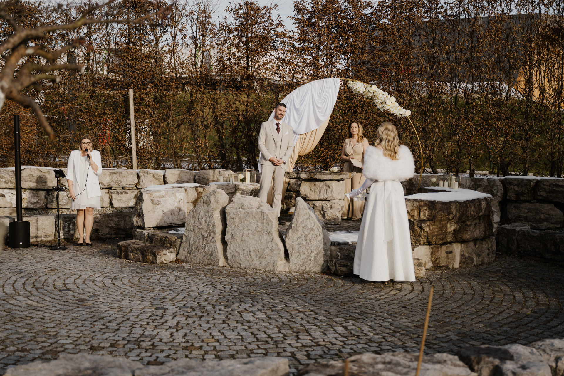 Eine winterliche Trauung im Freien, bei der vier in Weiß und Beige gekleidete Personen auf einer gepflasterten Fläche, umgeben von kahlen Bäumen und Steinstrukturen, unter einem geschmückten Bogen stehen.