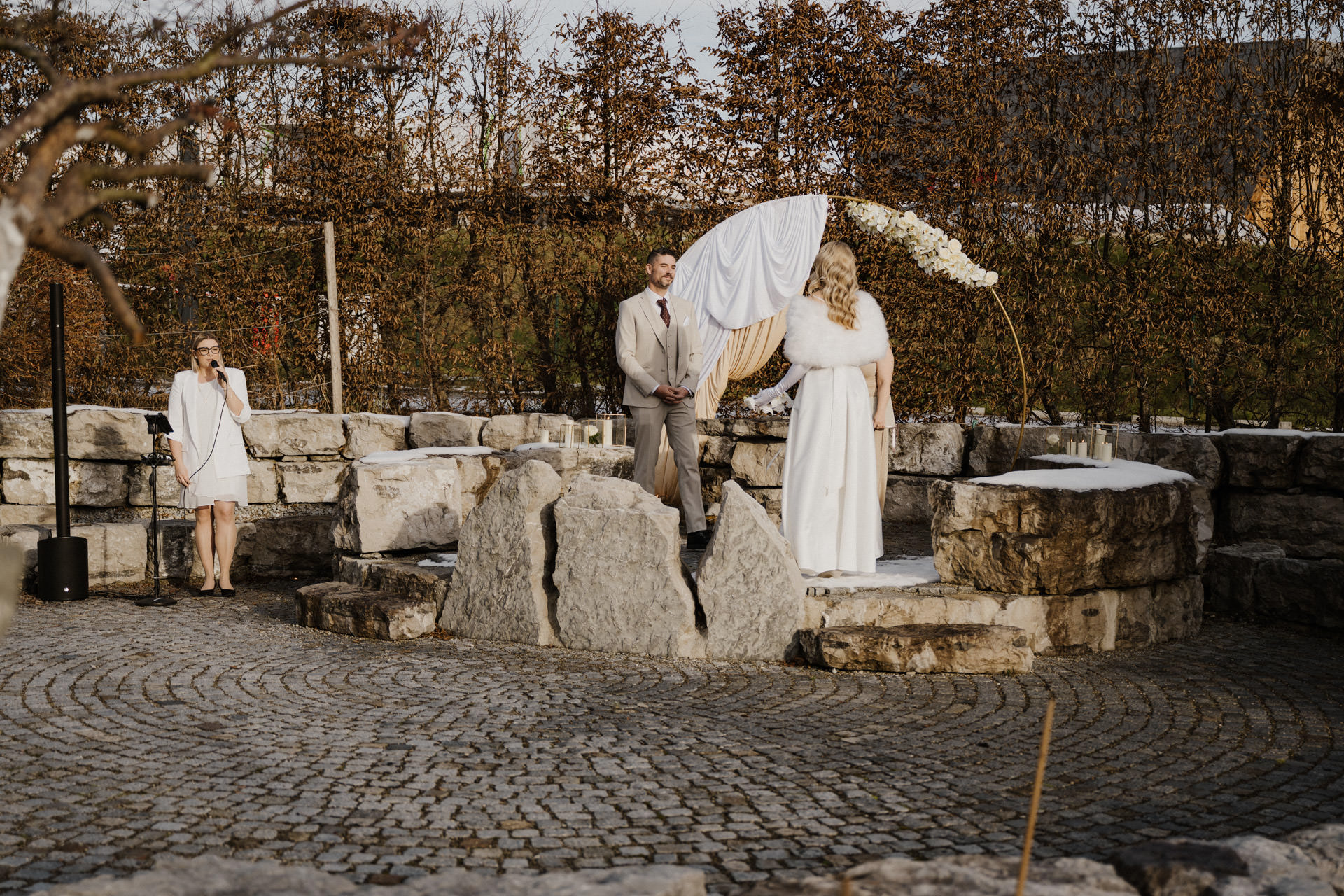 Ein weiß gekleidetes Brautpaar steht auf einem Steinpodest unter einem Blumenbogen im Freien. Links daneben steht eine Frau mit einem Mikrofon in der Hand, die möglicherweise die Trauung vornimmt. Der Boden und die Steine sind leicht mit Schnee bestäubt.