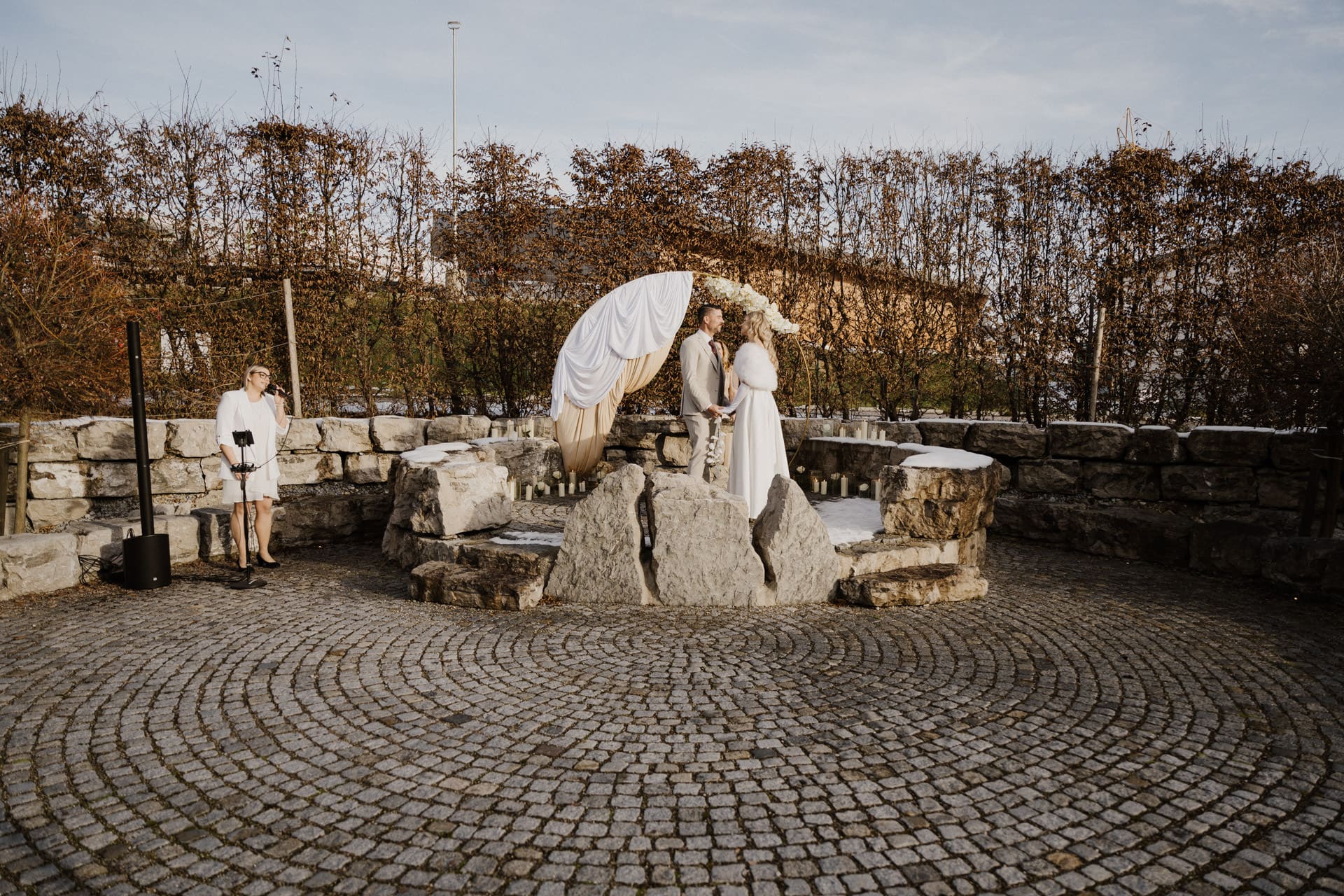 Zwei Bräute küssen sich unter einem mit weißem und beigem Stoff bespannten Bogen in einem Steinkreis im Freien. Links davon steht eine weiß gekleidete Person, die möglicherweise die Trauung vornimmt. Laublose Hecken und eine Steinmauer begrenzen die Szene.