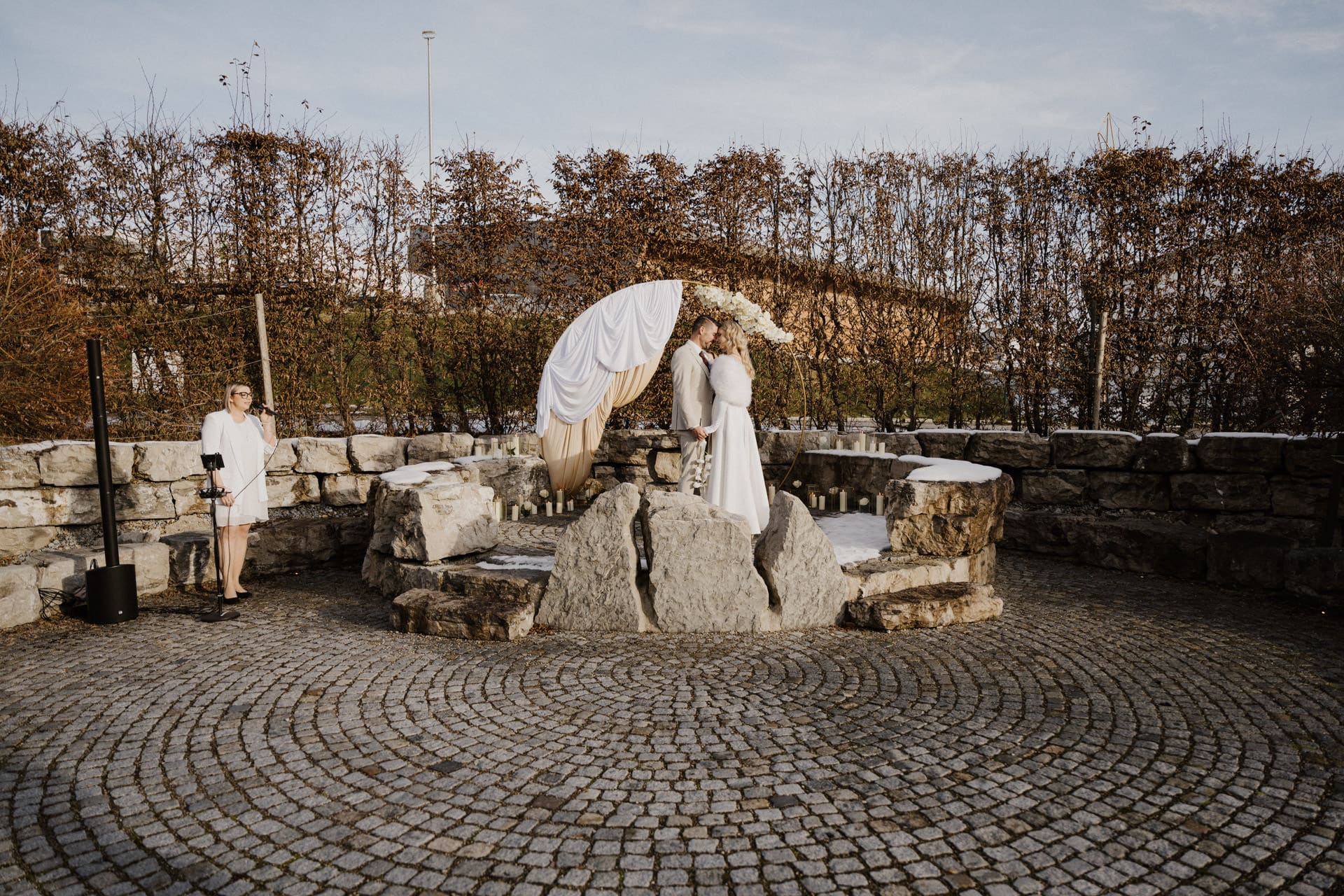 Ein weiß gekleidetes Paar steht unter einem Blumenbogen auf einem Steinpodest und scheint im Freien zu heiraten. Eine Person mit einer Kamera steht an der Seite. Kopfsteinpflaster und kahle Sträucher umgeben die Szene.